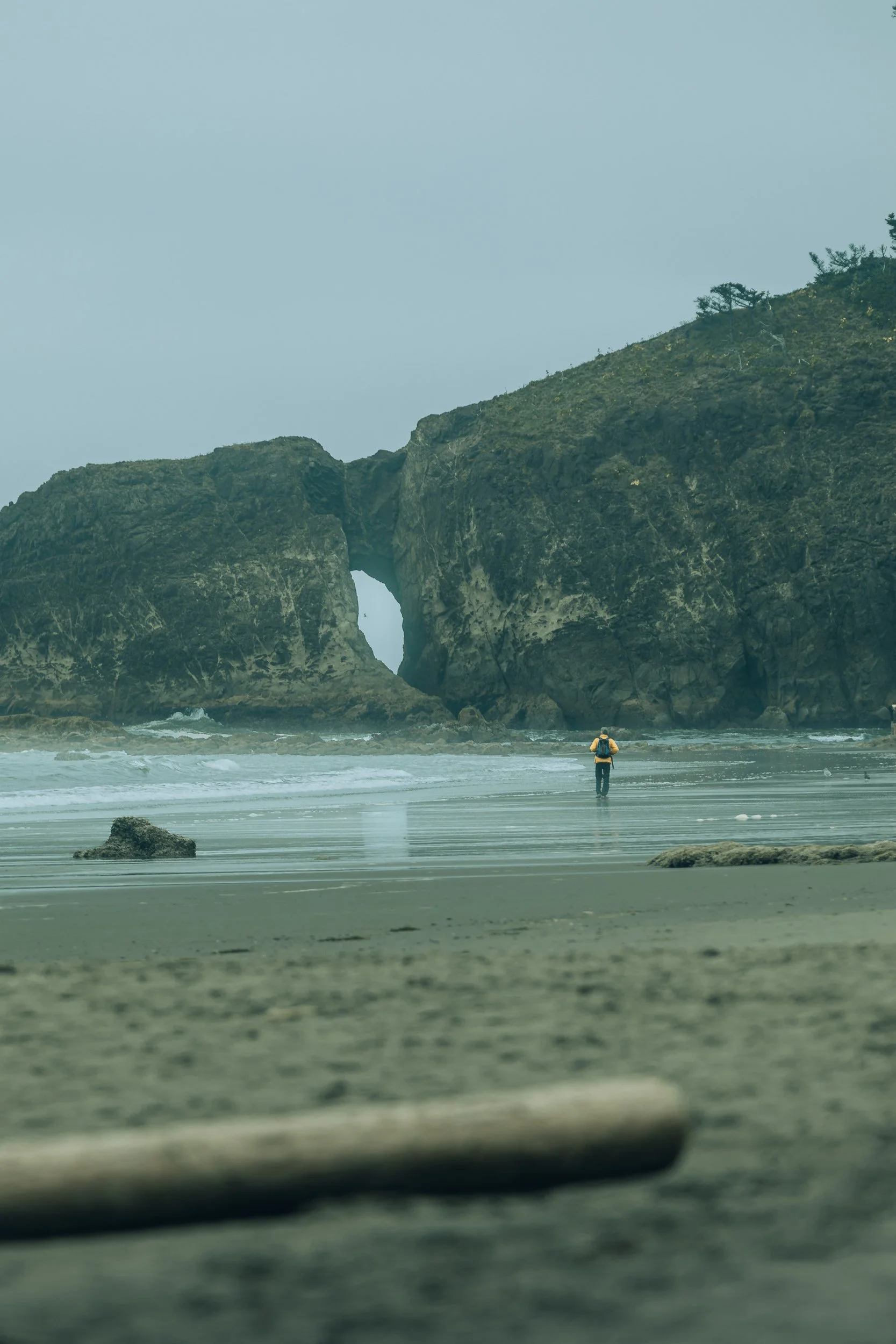 Person walking on wet sand toward a natural rock arch on the Olympic Peninsula coastline, Washington adventure travel photography by James Brasier