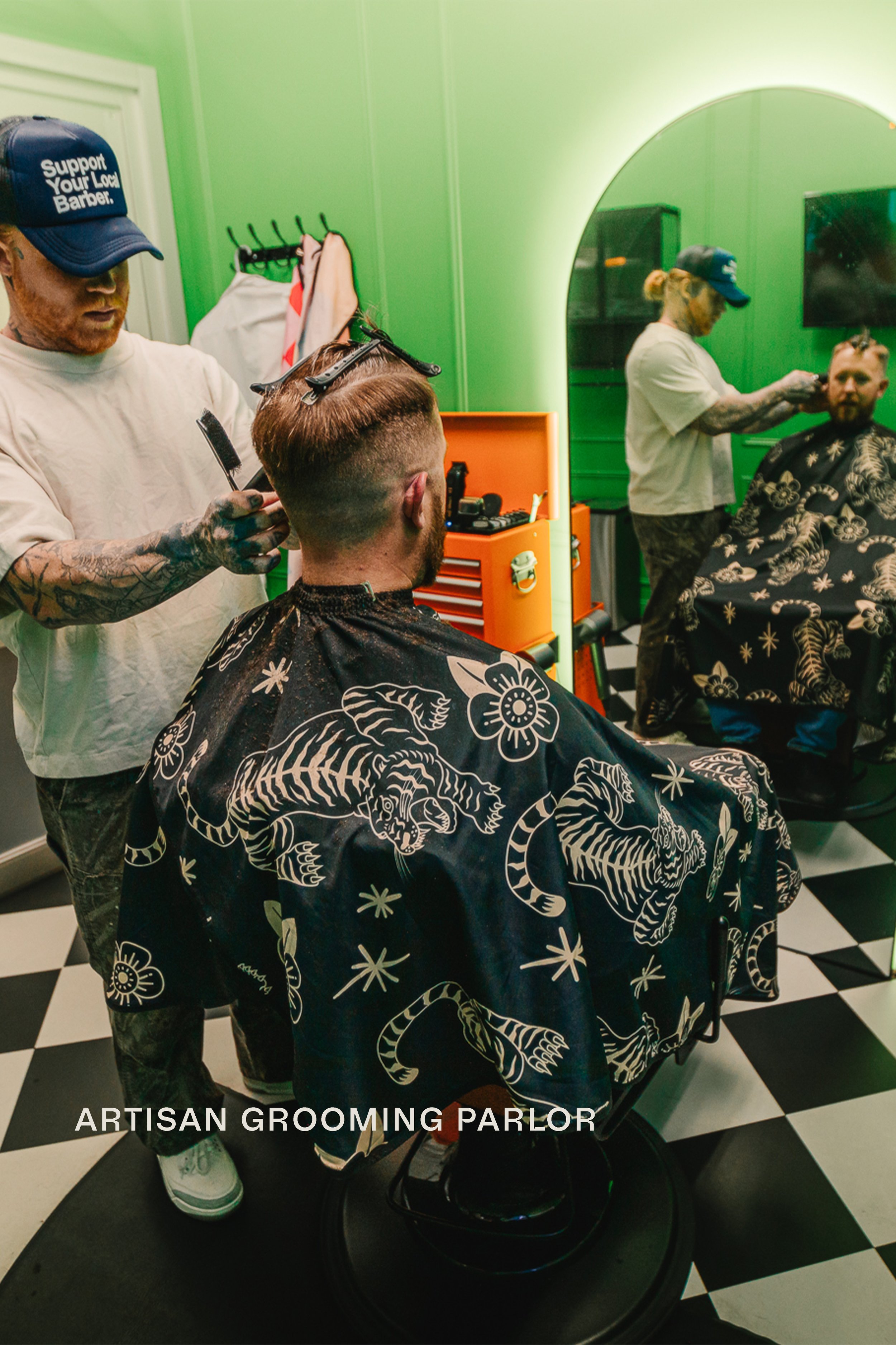 Barber cutting hair at Artisan Grooming Parlor with tiger print cape and mirror reflection against green walls and checkerboard floor, commercial photography by James Brasier