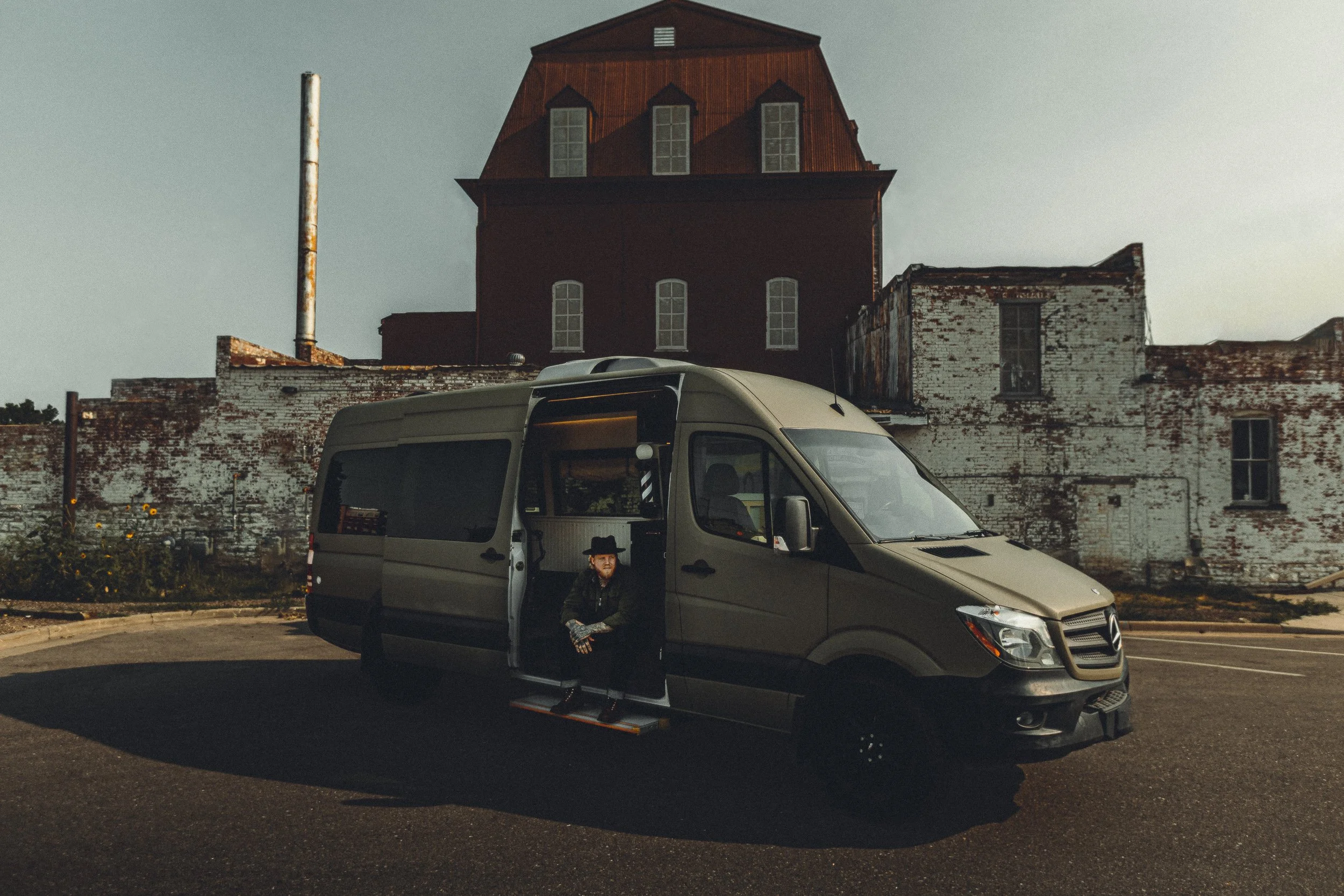 Nomad Mobile Barbershop owner sitting in the van doorway in front of an industrial building in Loveland Colorado, brand lifestyle photography by James Brasier