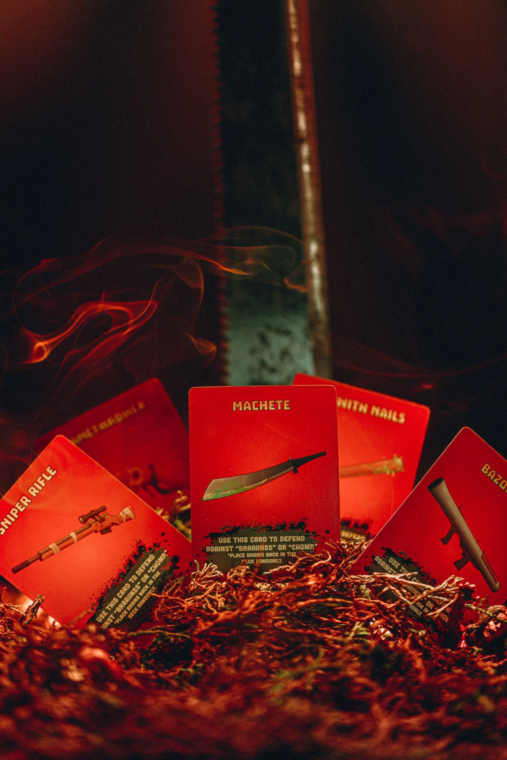 ZETA Survival weapon cards fanned with machete, sniper rifle, and bazooka under red lighting with smoke, product photography by James Brasier