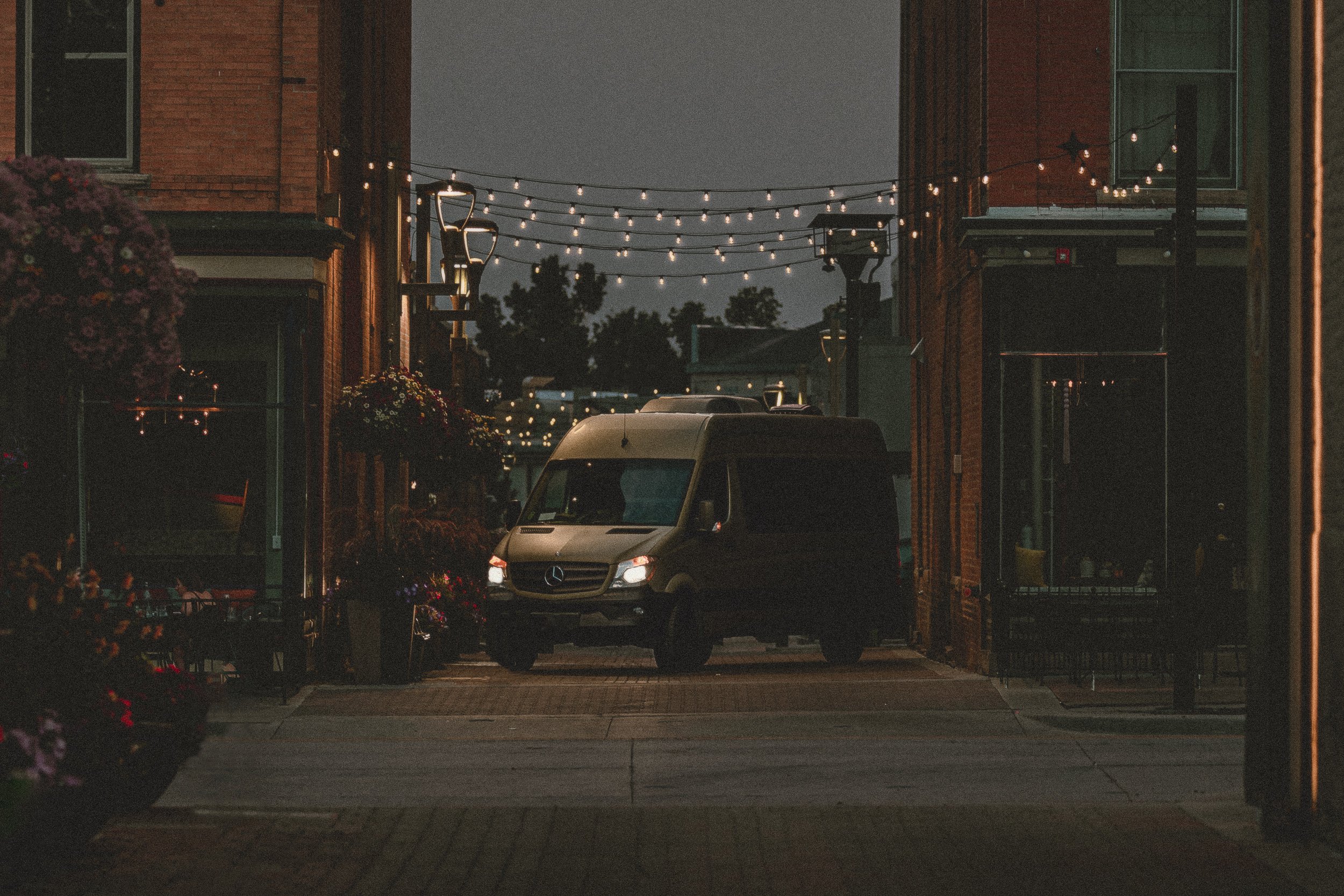 Nomad Mobile Barbershop van driving through a downtown alley at dusk with headlights on and string lights overhead in Fort Collins Colorado, commercial photography by James Brasier