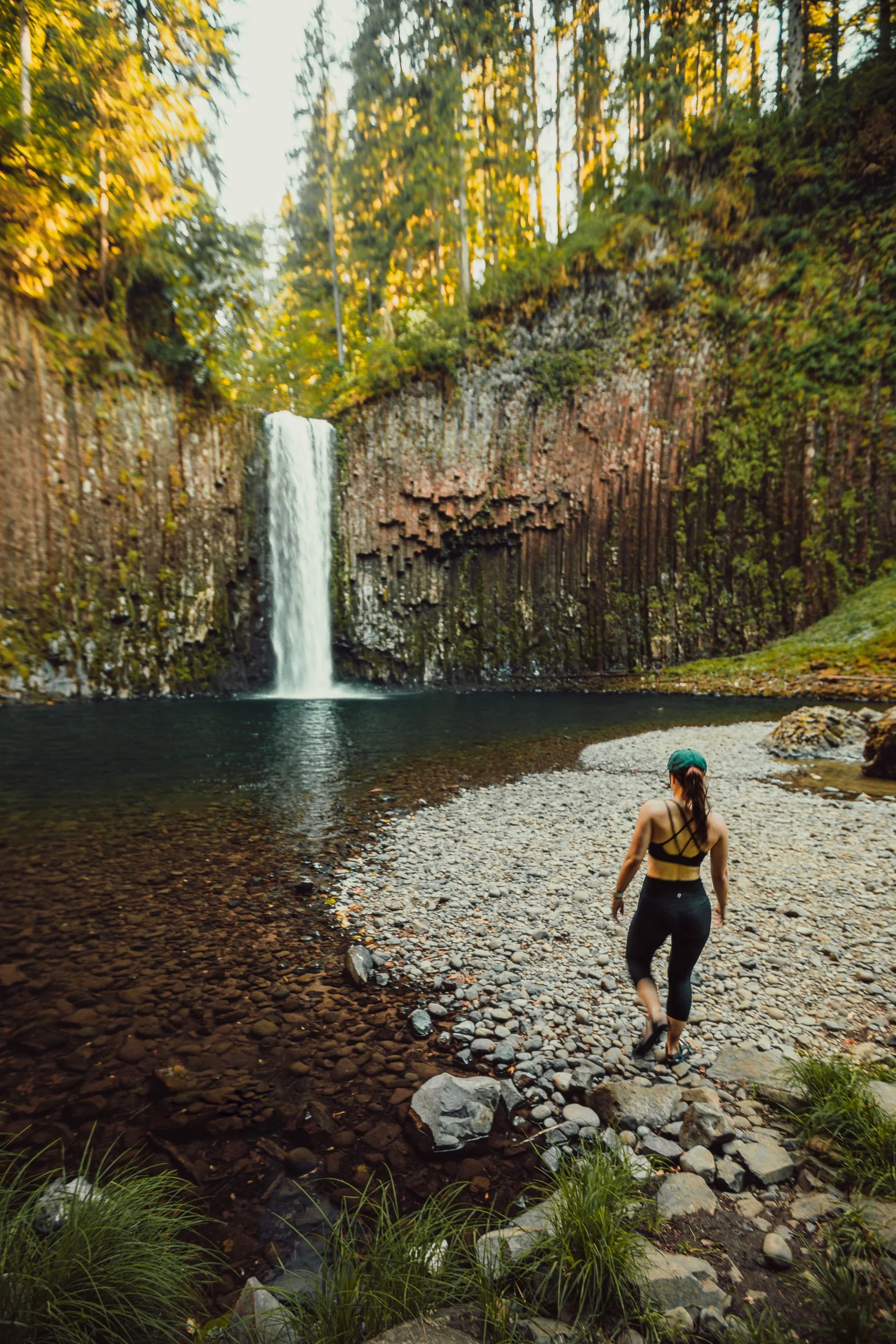 Person walking toward a waterfall along the rocky shoreline beneath basalt column walls in an Oregon rainforest, adventure photography by James Brasier