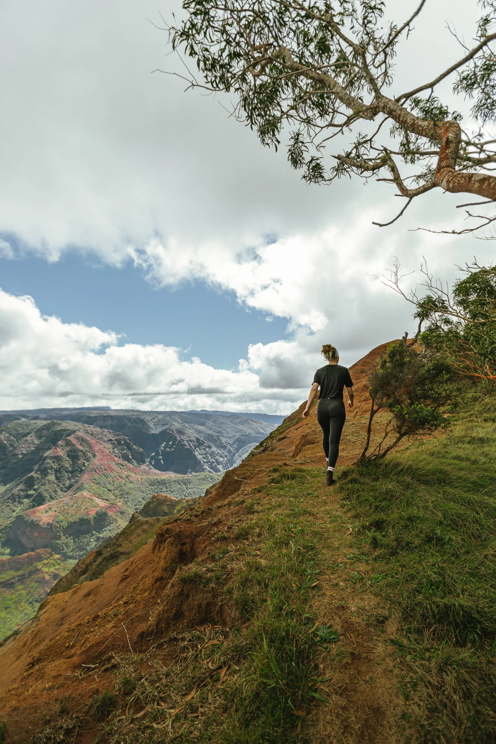Hiker walking along a red dirt ridge above Waimea Canyon with layered canyon walls in the distance, Kauai Hawaii adventure photography by James Brasier
