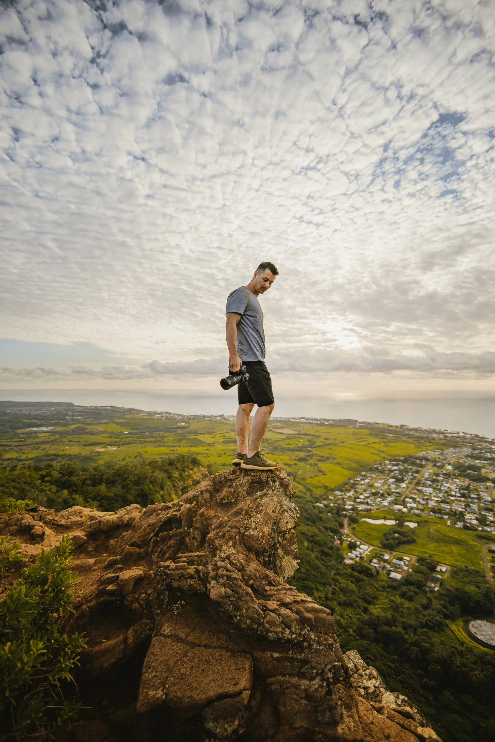 Person standing on the summit of Sleeping Giant with camera overlooking green farmland and coastline, Kauai Hawaii outdoor photography by James Brasier