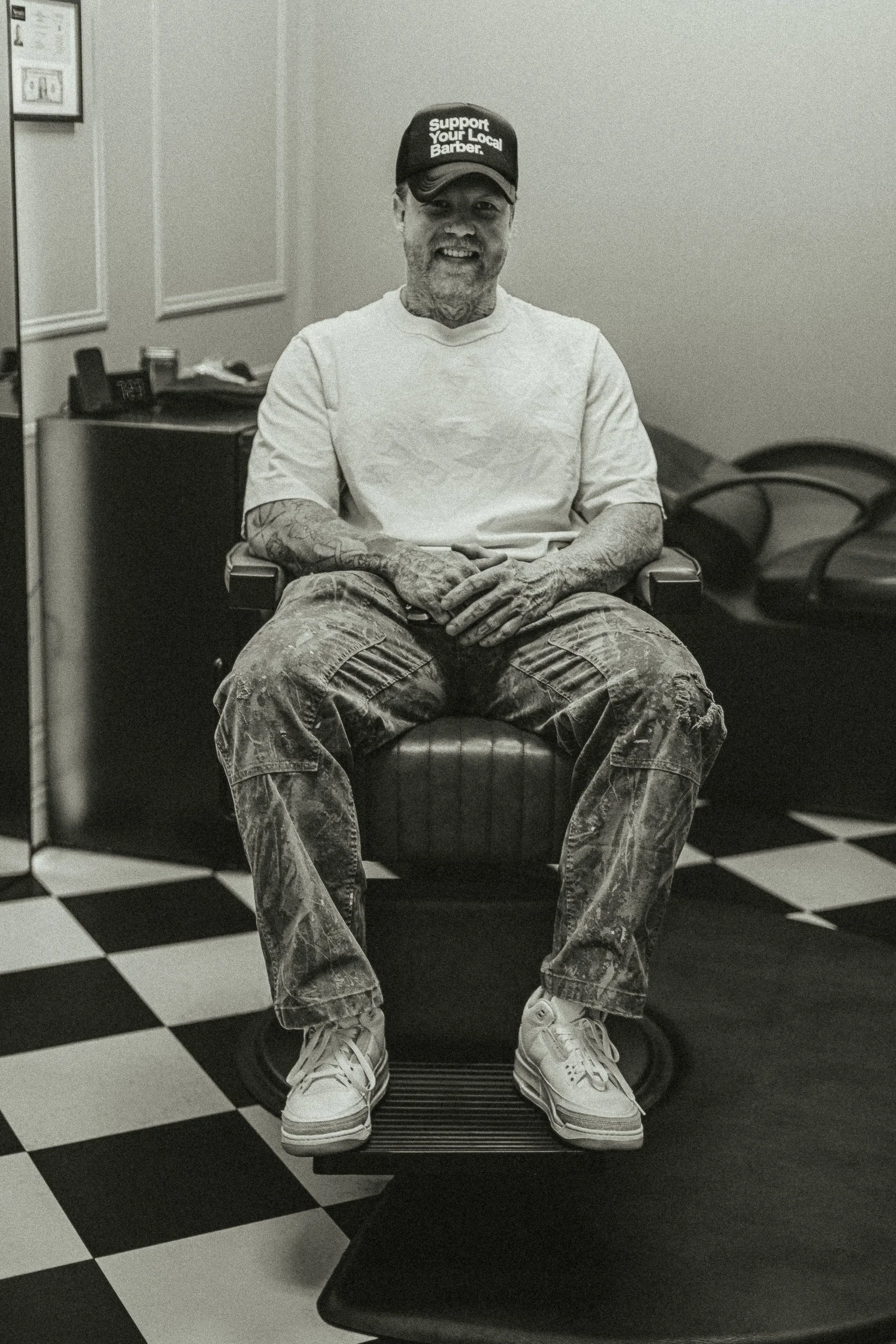 Artisan Grooming Parlor owner sitting in the barber chair on a checkerboard floor in paint-stained pants in black and white, brand portrait photography by James Brasier