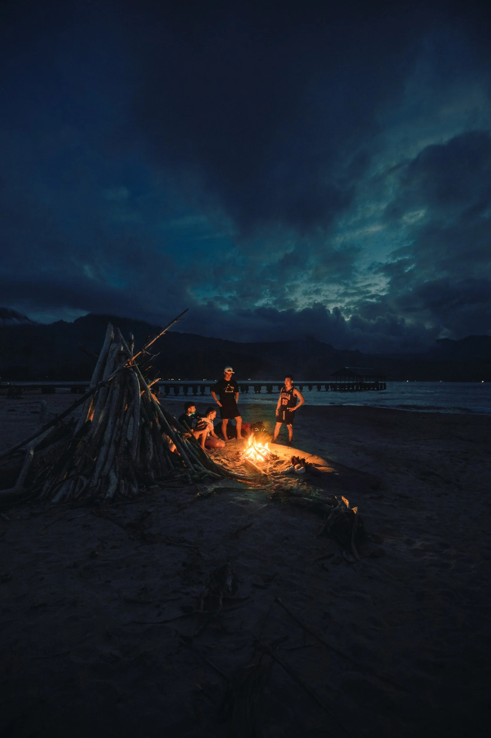 Group of friends gathered around a driftwood bonfire on the beach at Hanalei Bay with mountains and pier in the background at dusk, Kauai Hawaii adventure lifestyle photography by James Brasier