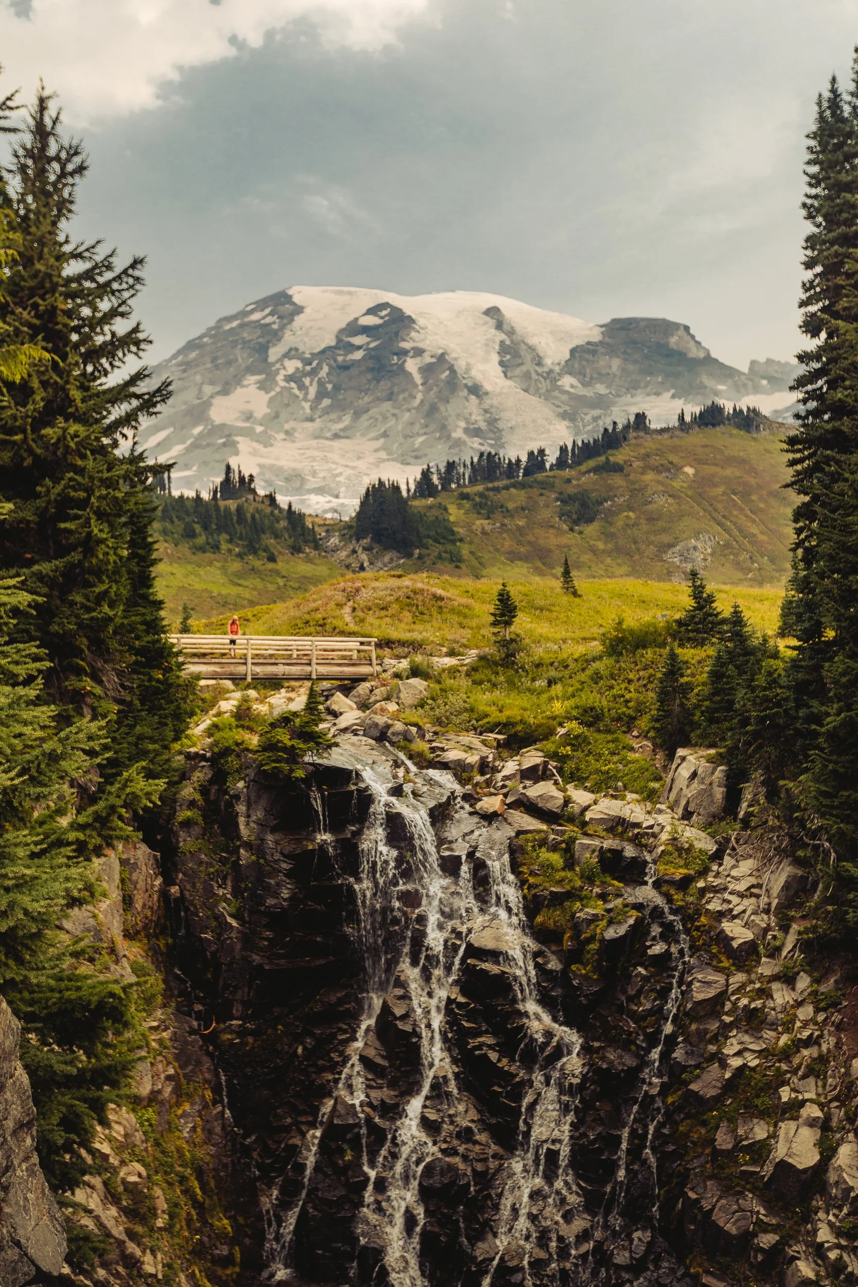 Waterfall cascading over rock face with hiker on bridge and Mount Rainier glaciers in the background, Mount Rainier National Park photography by James Brasier