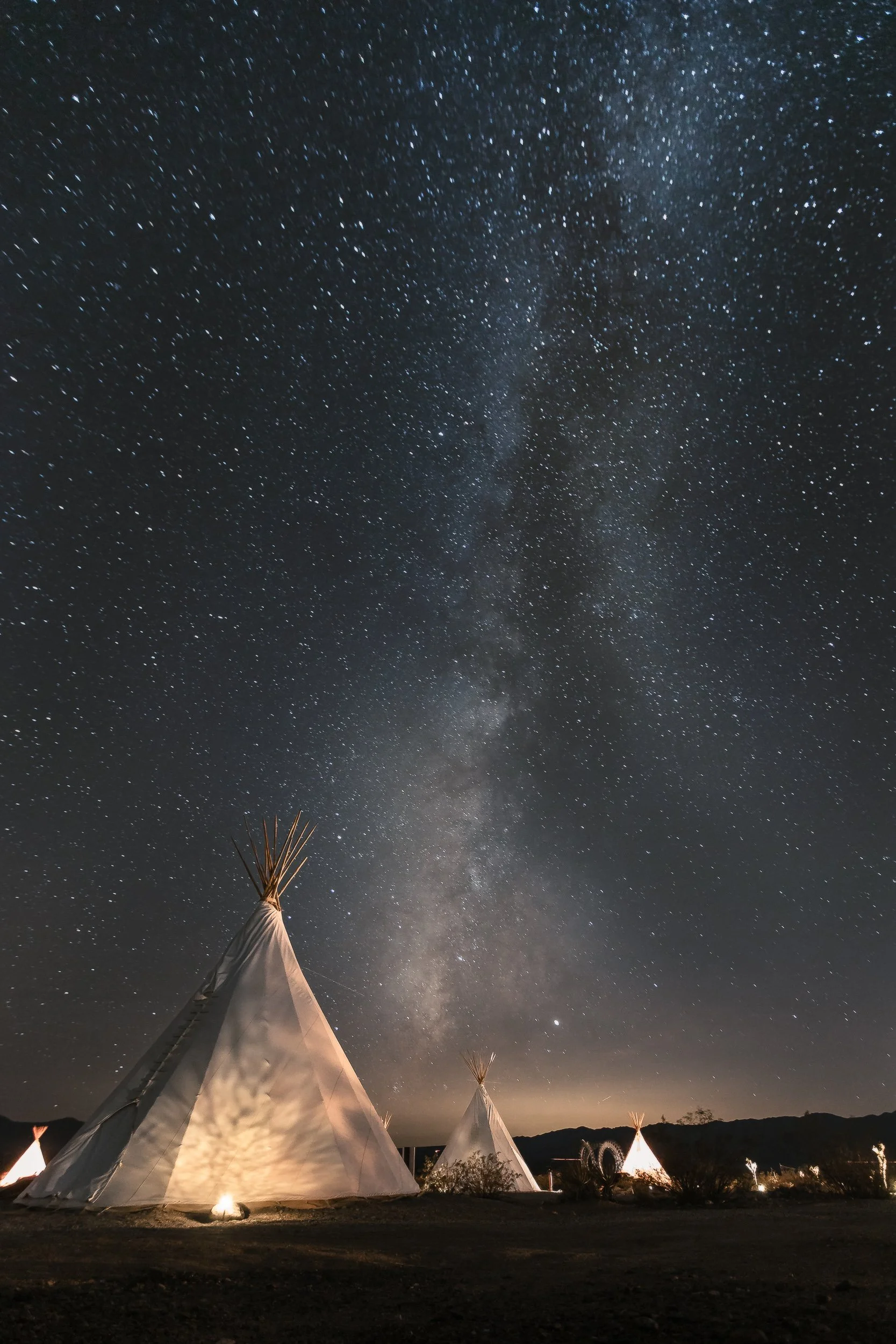 Teepees beneath the Milky Way with campfire glow at a remote desert camp in the Mojave, astrophotography by James Brasier