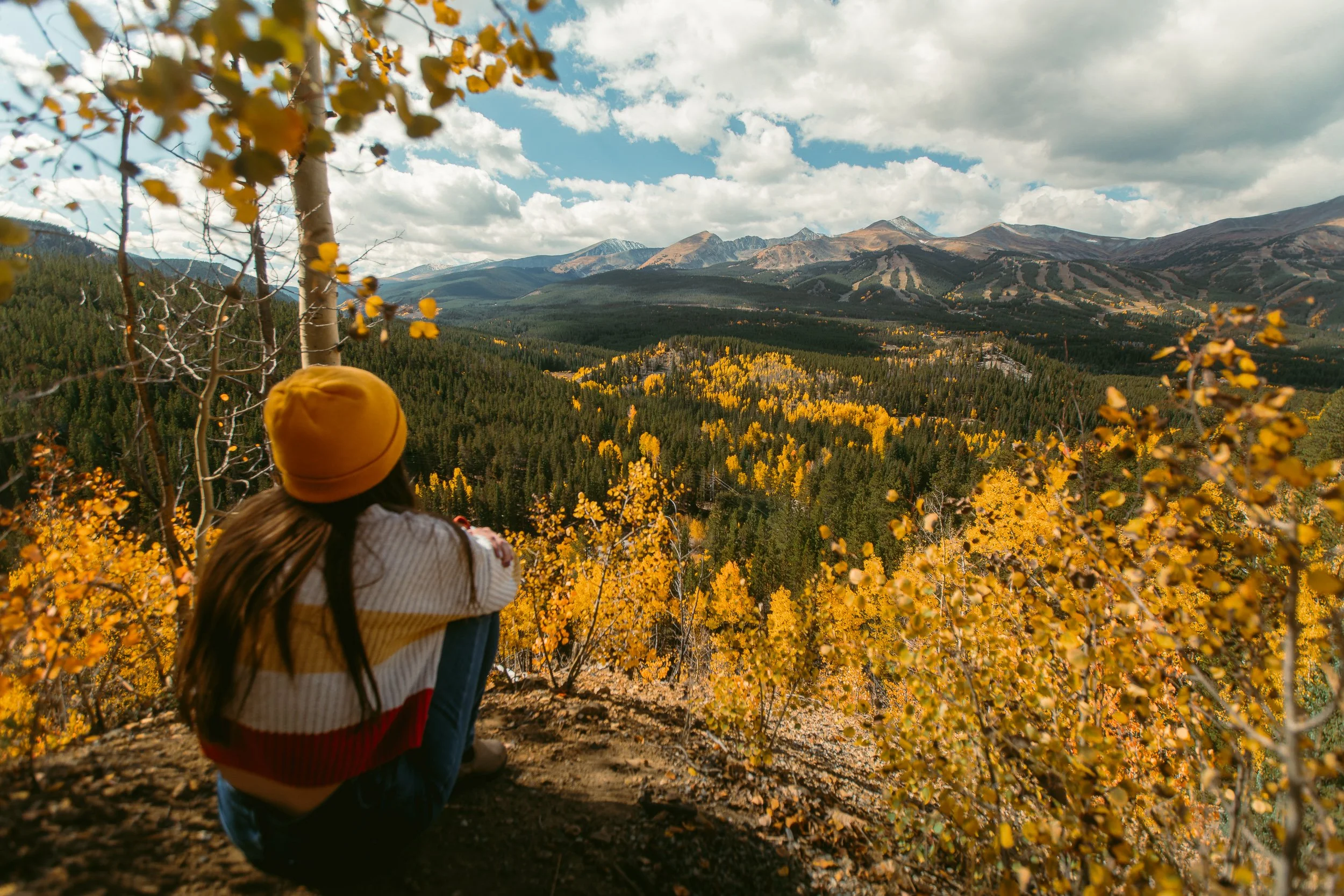 Person in yellow beanie sitting among golden fall aspens overlooking mountain valley near Breckenridge Colorado, autumn landscape photography by James Brasier