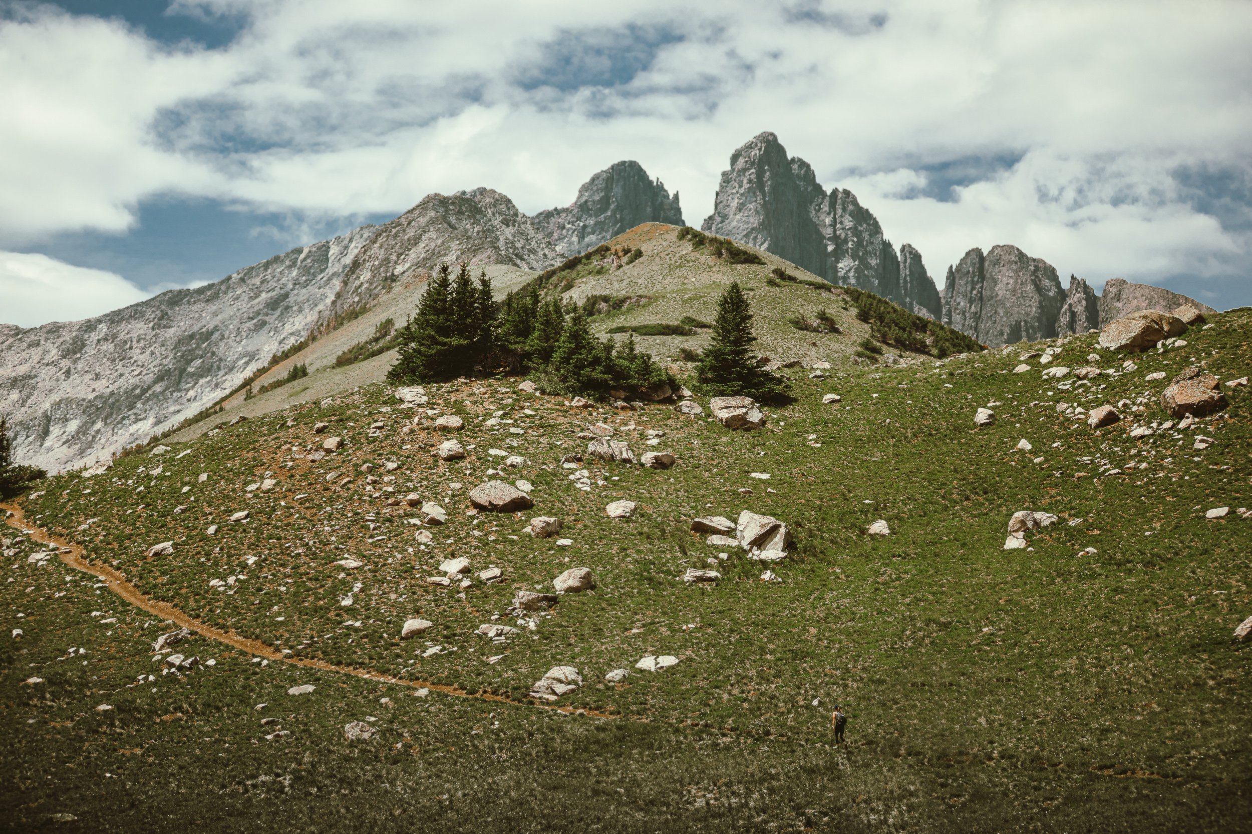 Hiker on trail through alpine meadow with jagged granite peaks at Michigan Lakes in the Colorado high country, landscape photography by James Brasier