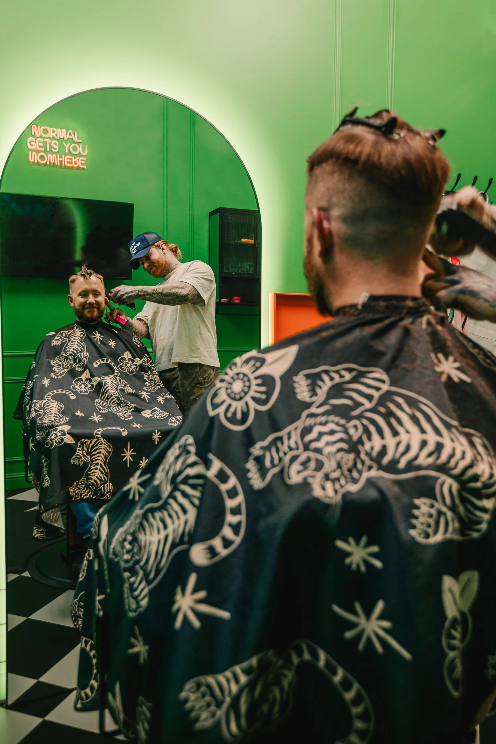 Mirror reflection of barber cutting hair with neon sign and tiger print cape in the foreground at Artisan Grooming Parlor, commercial photography by James Brasier