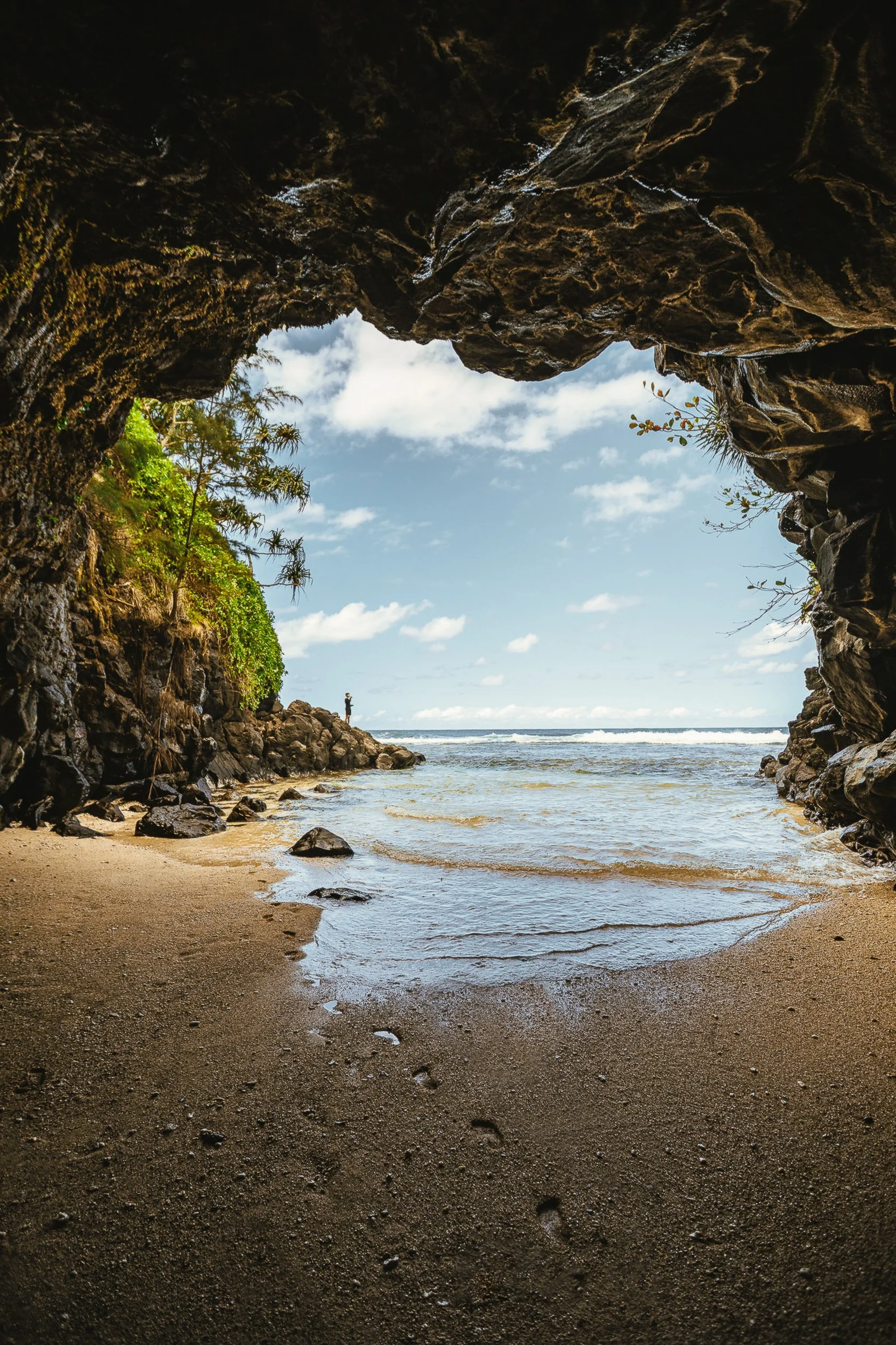 View from inside a lava rock sea cave looking out to ocean waves and a sandy beach on the Kauai coastline, Hawaii adventure travel photography by James Brasier