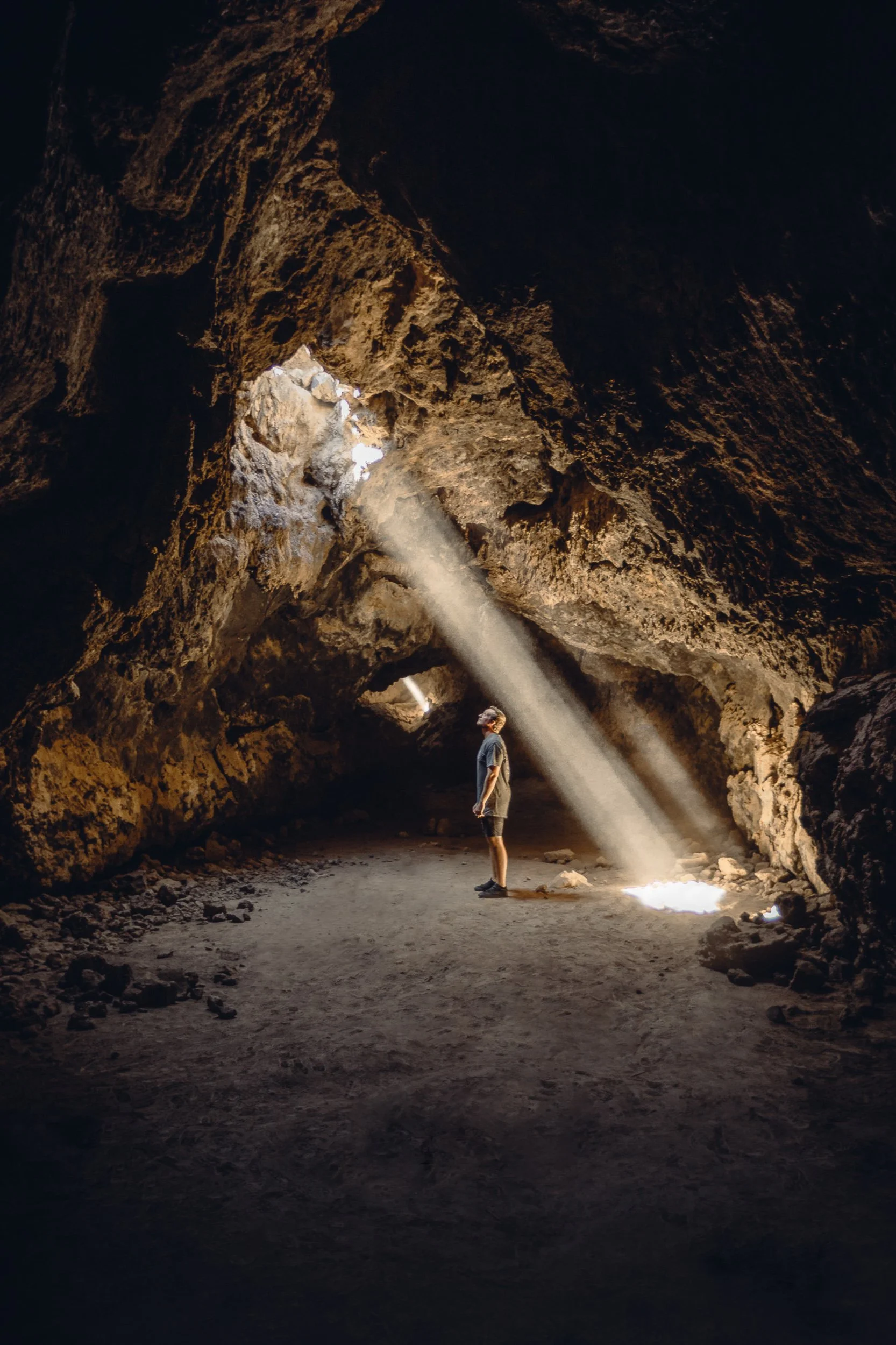 Person standing in a Mojave Desert lava tube with a beam of light streaming through the cave ceiling, adventure photography by James Brasier