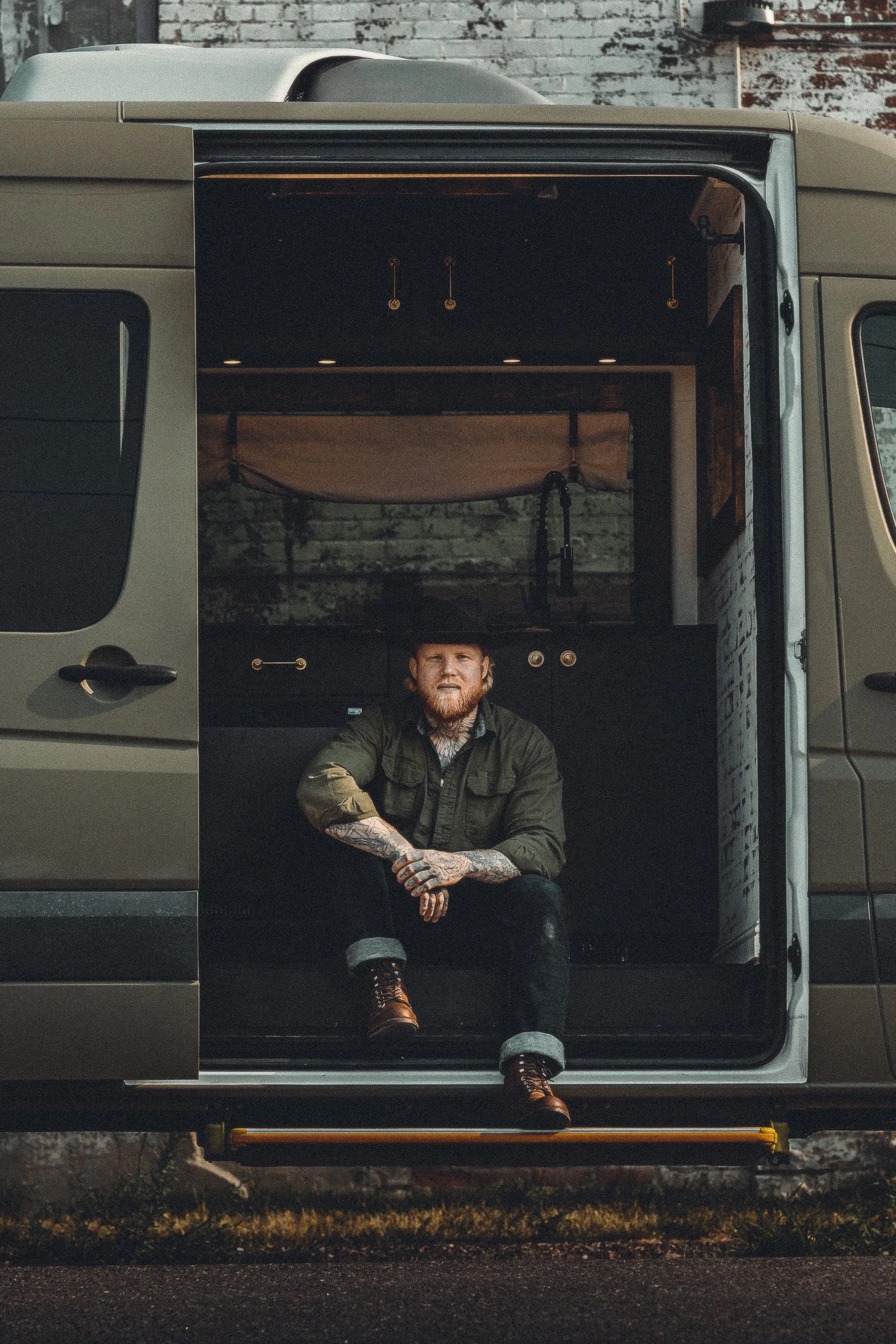 Nomad Mobile Barbershop owner sitting in the van doorway with tattoos and boots visible and the interior glowing behind him, brand portrait photography by James Brasier