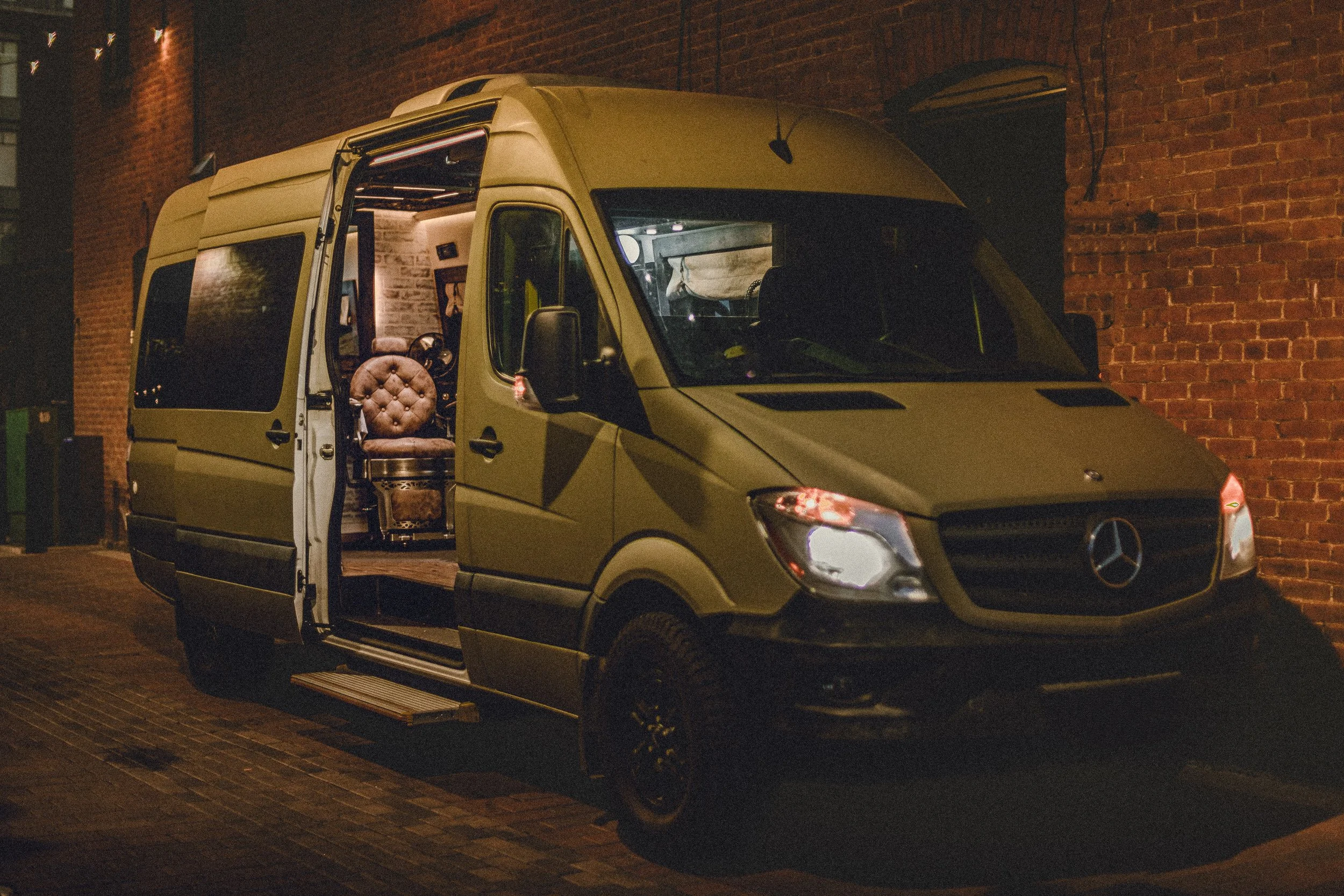 Nomad Mobile Barbershop van with side door open showing barber chair and headlights glowing in a brick alley in Fort Collins Colorado, commercial photography by James Brasier