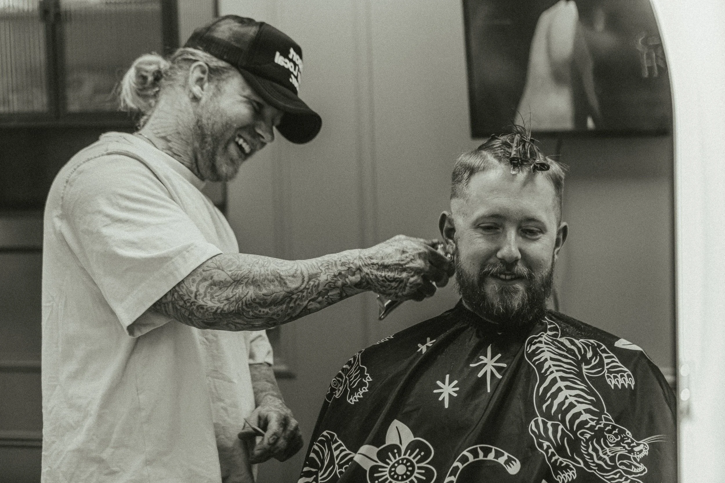 Barber and client laughing during a haircut at Artisan Grooming Parlor in black and white, candid commercial photography by James Brasier