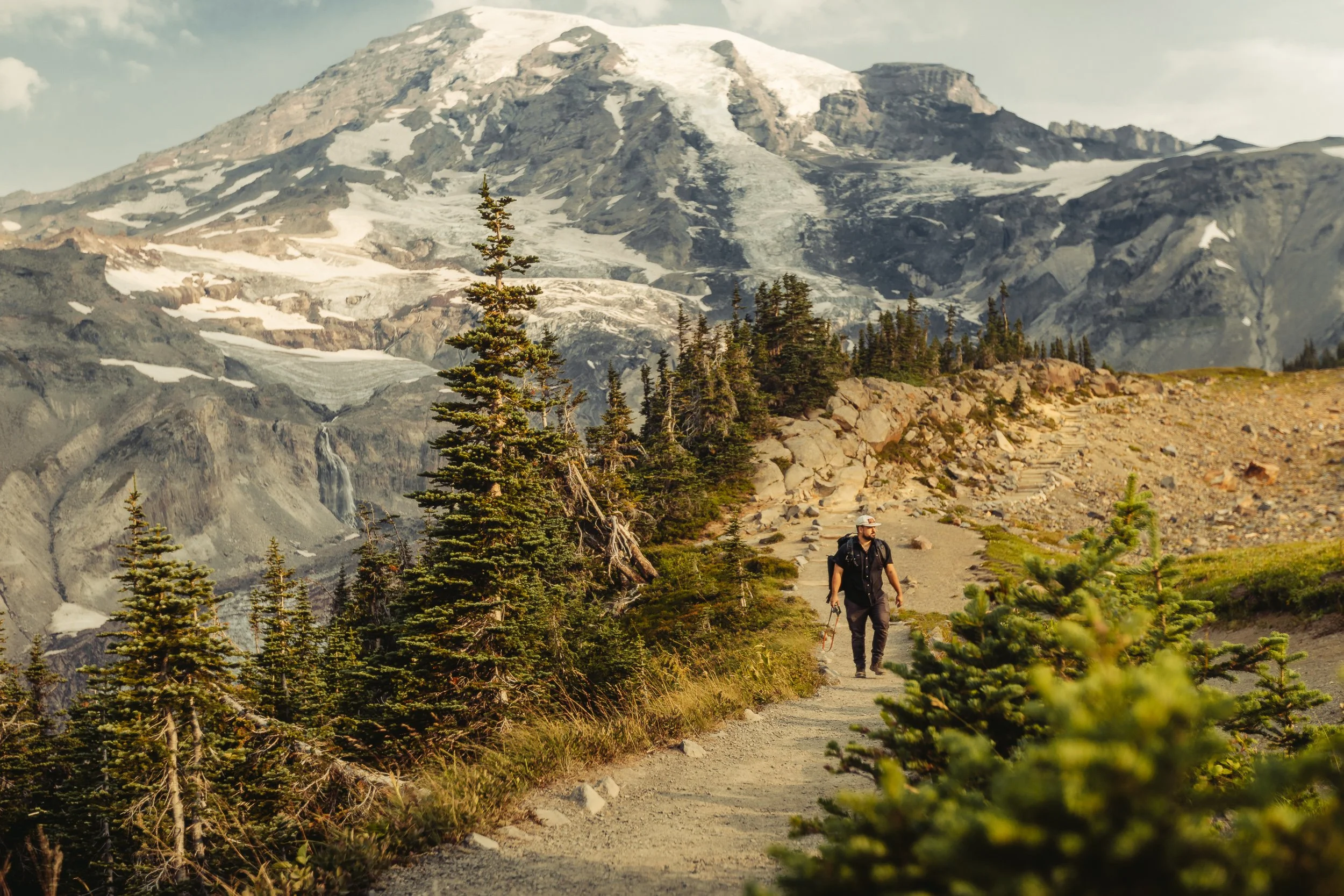 Hiker walking along alpine trail with Mount Rainier and glacial waterfall behind, outdoor adventure photography by James Brasier