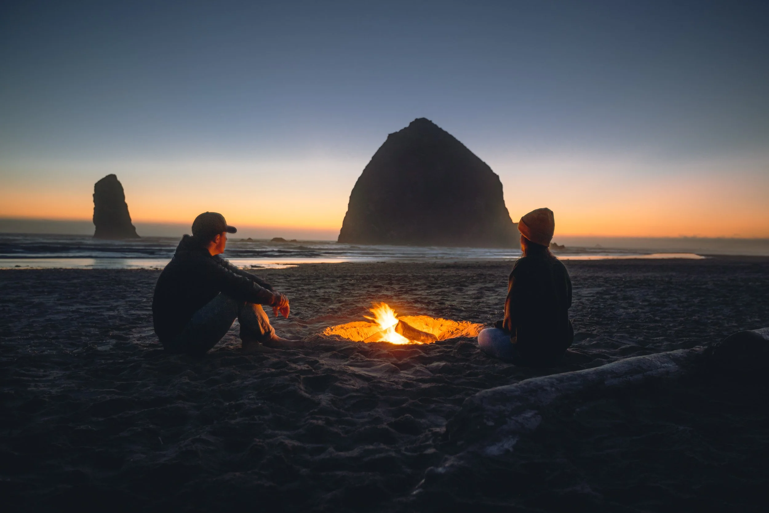 Two people sitting by a beach bonfire with Haystack Rock silhouetted at dusk at Cannon Beach Oregon, outdoor lifestyle photography by James Brasier