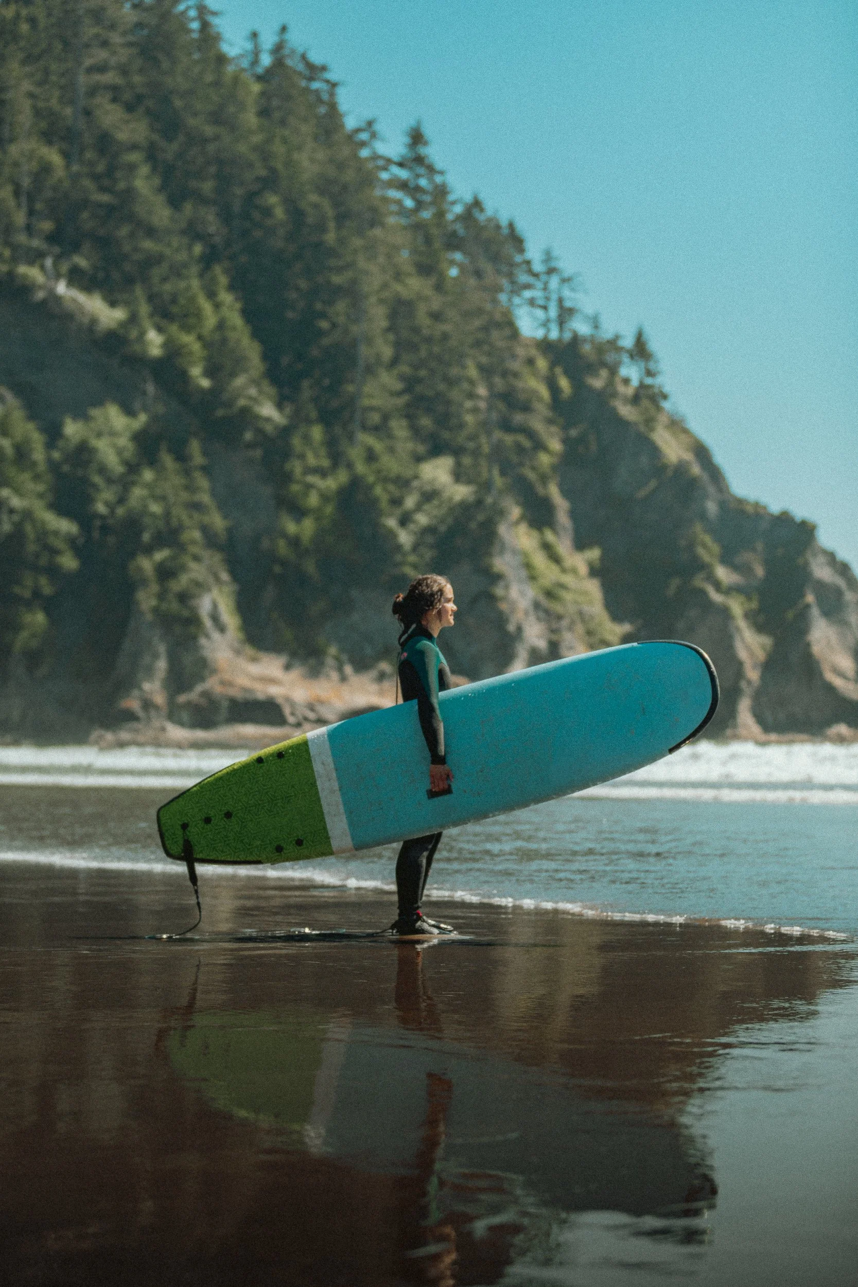 Surfer carrying a longboard on the beach with forested sea cliffs on the Oregon Coast, surf photography by James Brasier