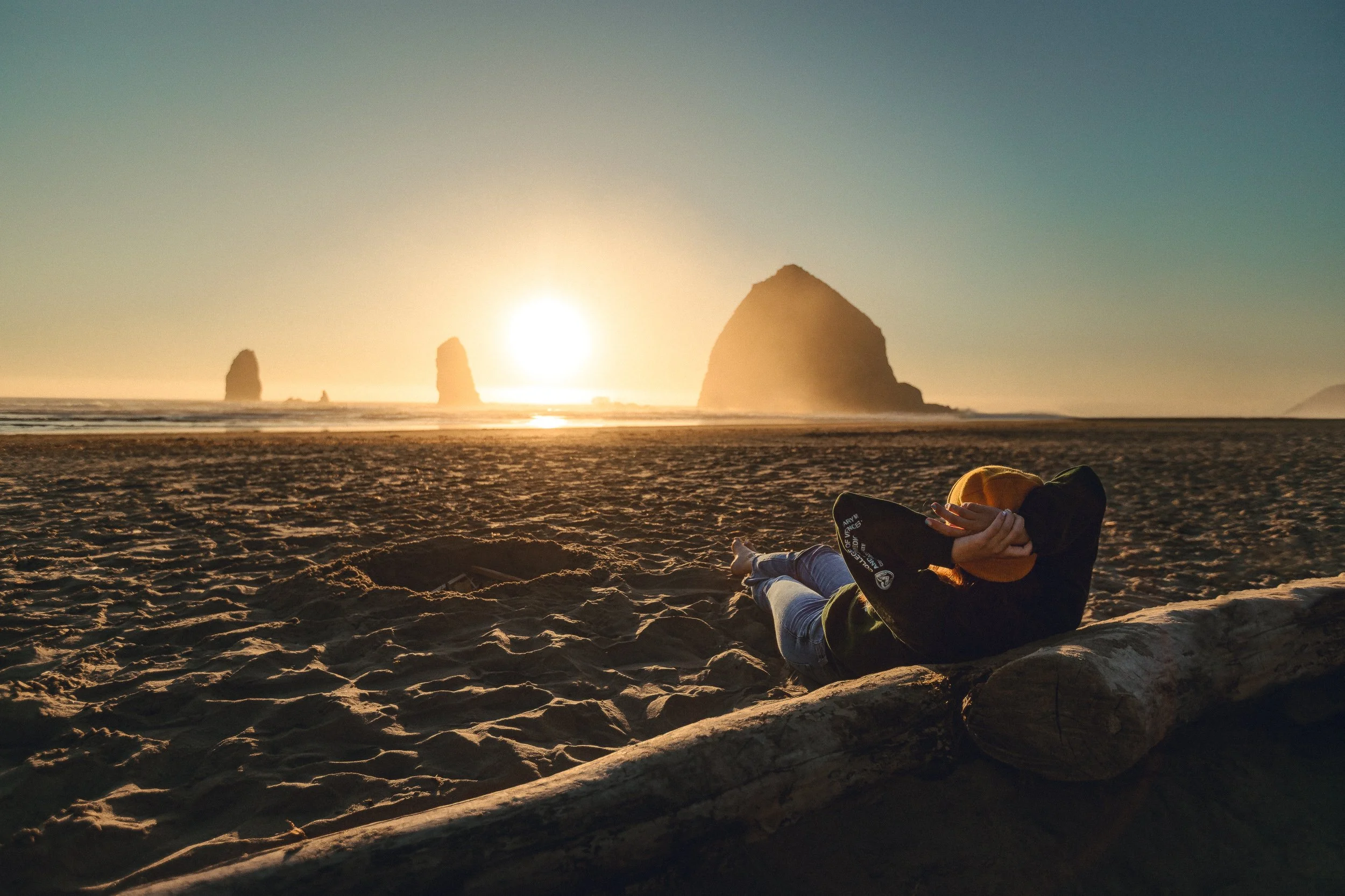 Person relaxing on driftwood watching the sunset behind Haystack Rock at Cannon Beach Oregon, Oregon Coast photography by James Brasier