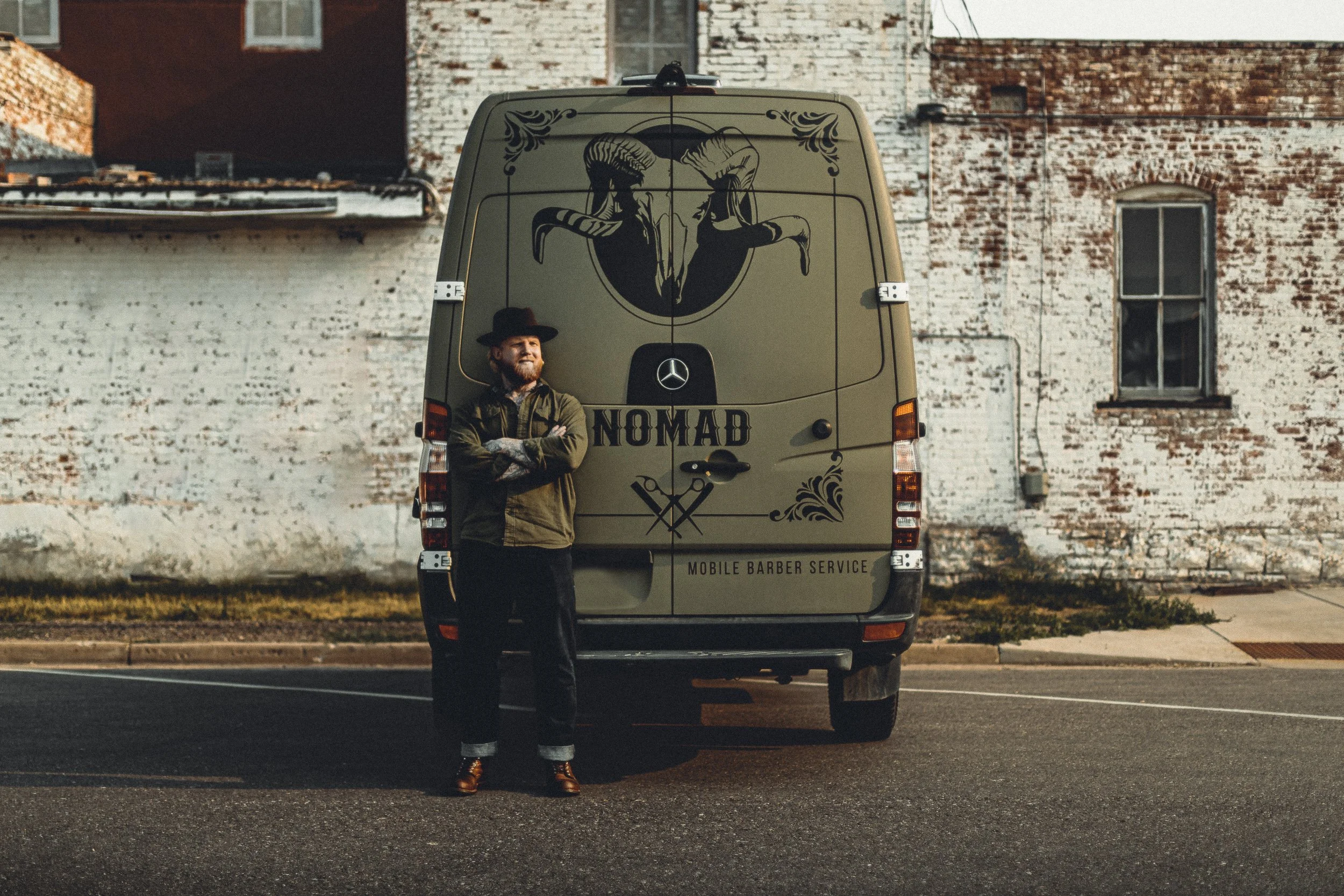Nomad Mobile Barbershop owner leaning against the rear of the van with arms crossed and full brand graphics visible, brand identity photography by James Brasier