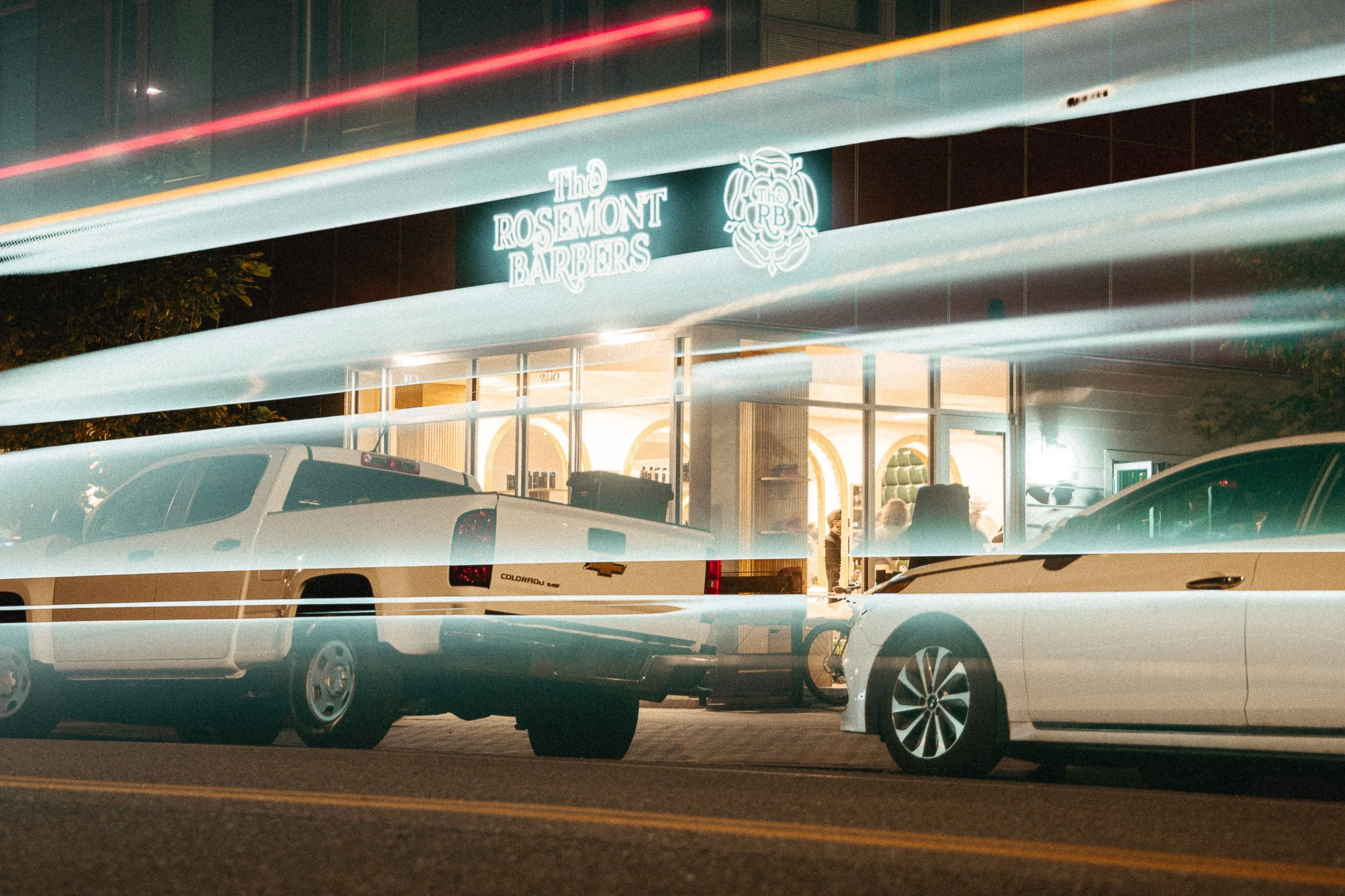 The Rosemont Barbers storefront at night with neon signage and car light trails in Denver's RiNo district, commercial photography by James Brasier