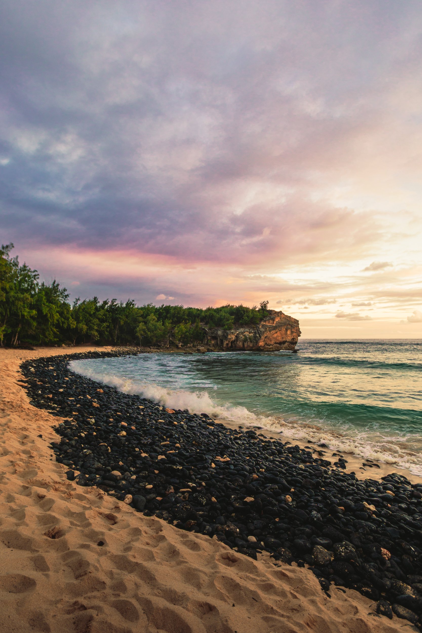 Black volcanic rock and sand beach curving toward sea cliffs at sunset with purple and pink skies at Shipwreck Beach, Kauai Hawaii coastal photography by James Brasier