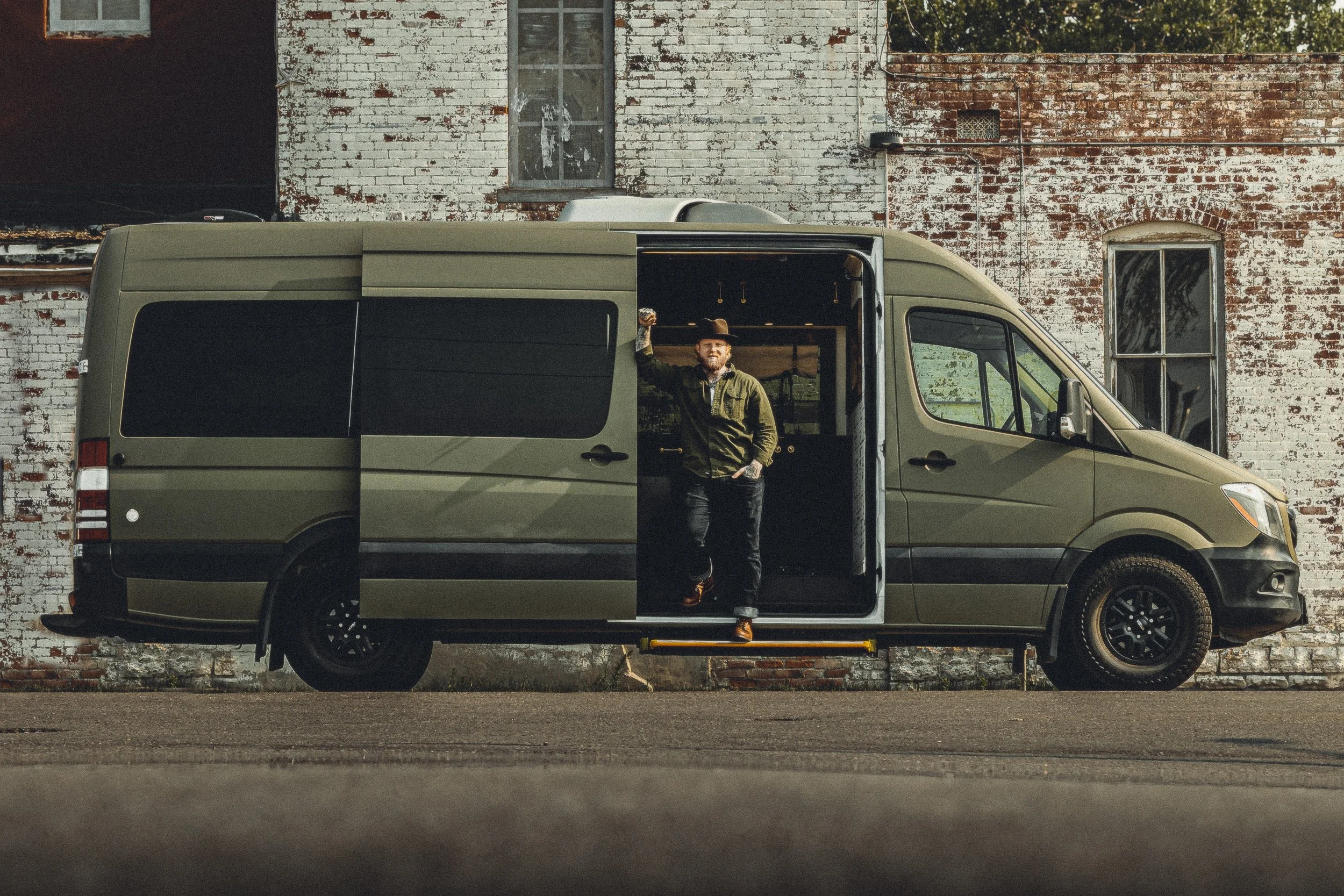 Nomad Mobile Barbershop owner standing in the side door of the van with full vehicle profile and brick building in Loveland Colorado, commercial photography by James Brasier