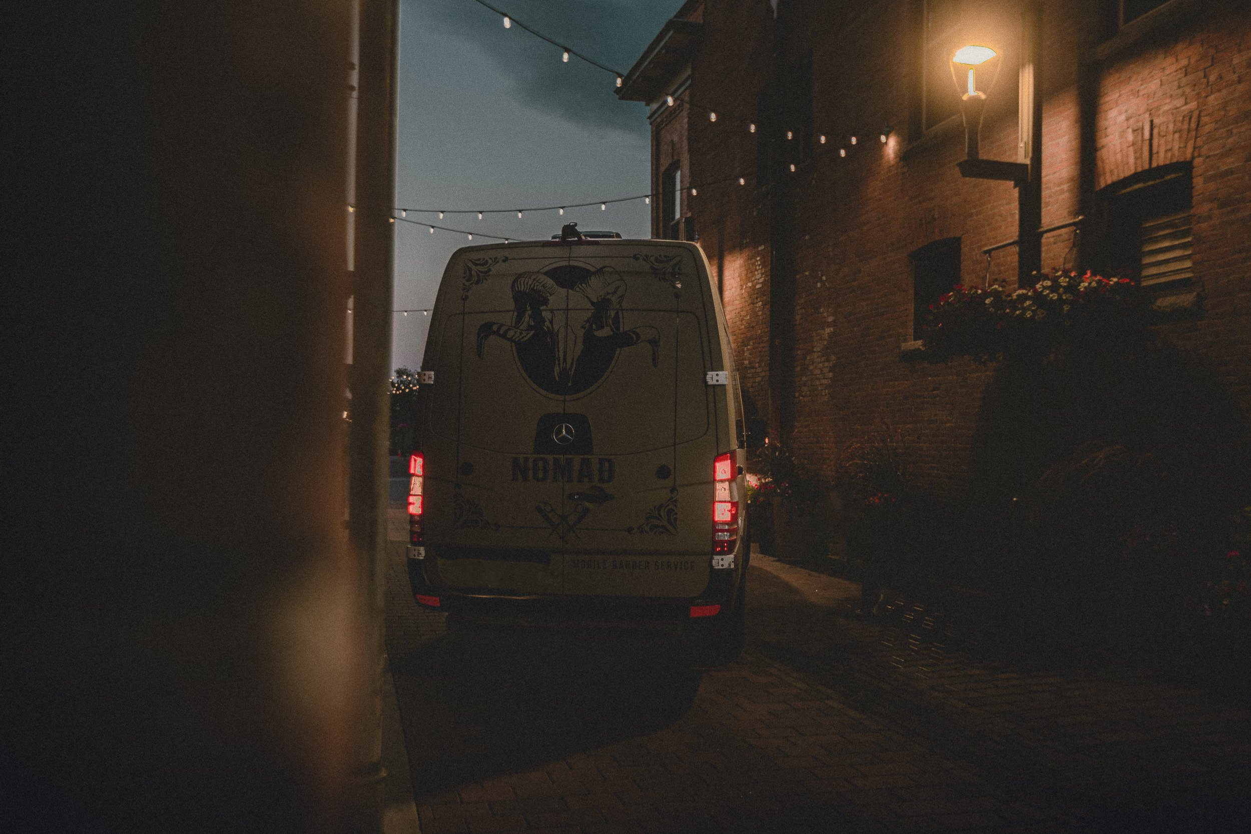 Nomad Mobile Barbershop van rear with brake lights and branding visible in a brick alley with string lights in Fort Collins Colorado, brand photography by James Brasier