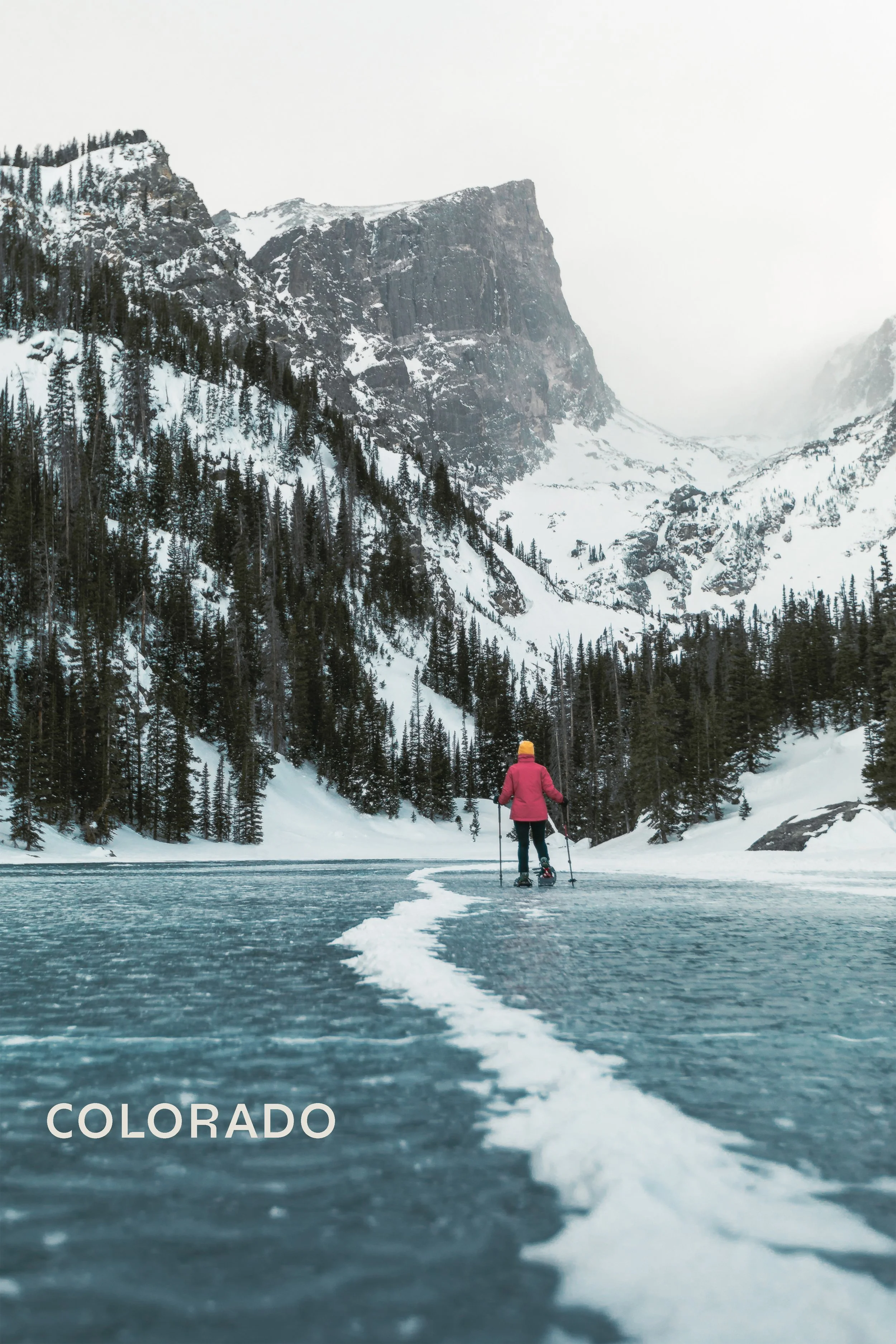 Hiker standing on frozen alpine lake beneath snow-covered peaks in Rocky Mountain National Park, Colorado landscape photography by James Brasier