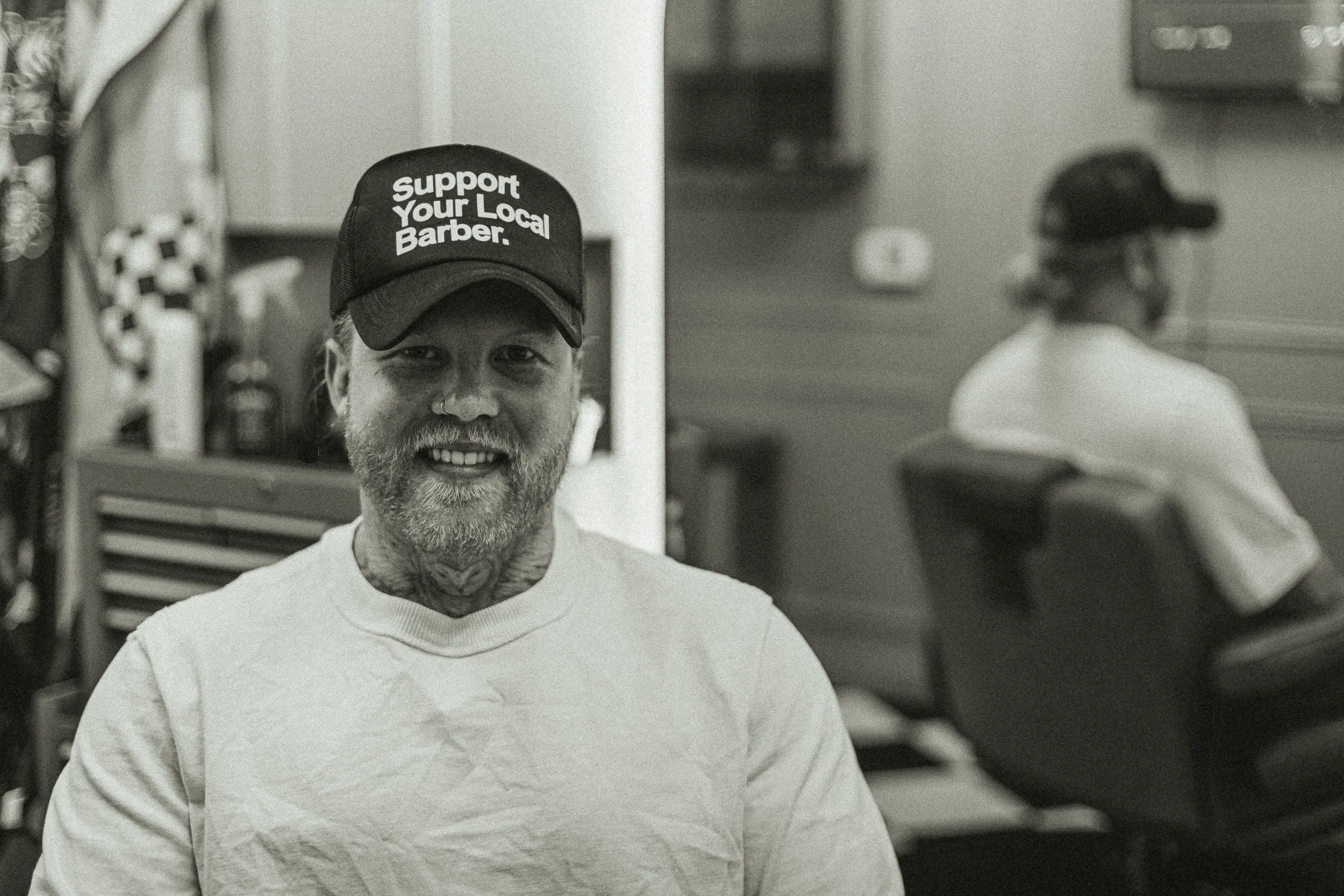 Artisan Grooming Parlor owner smiling in the barbershop wearing a Support Your Local Barber hat, commercial portrait photography by James Brasier