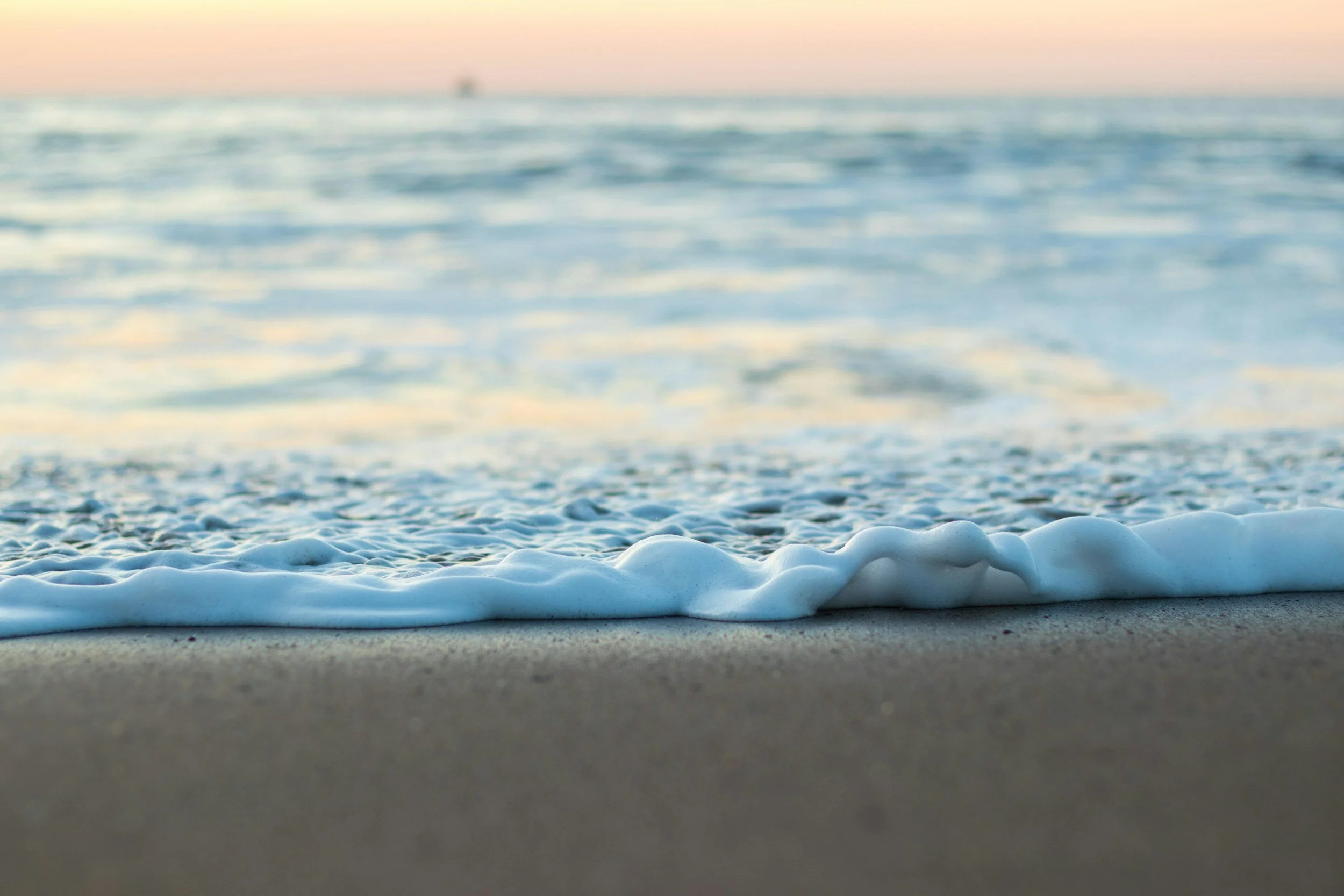 Close-up of ocean waves lapping onto sandy beach at sunset with blurred horizon and sky in background.