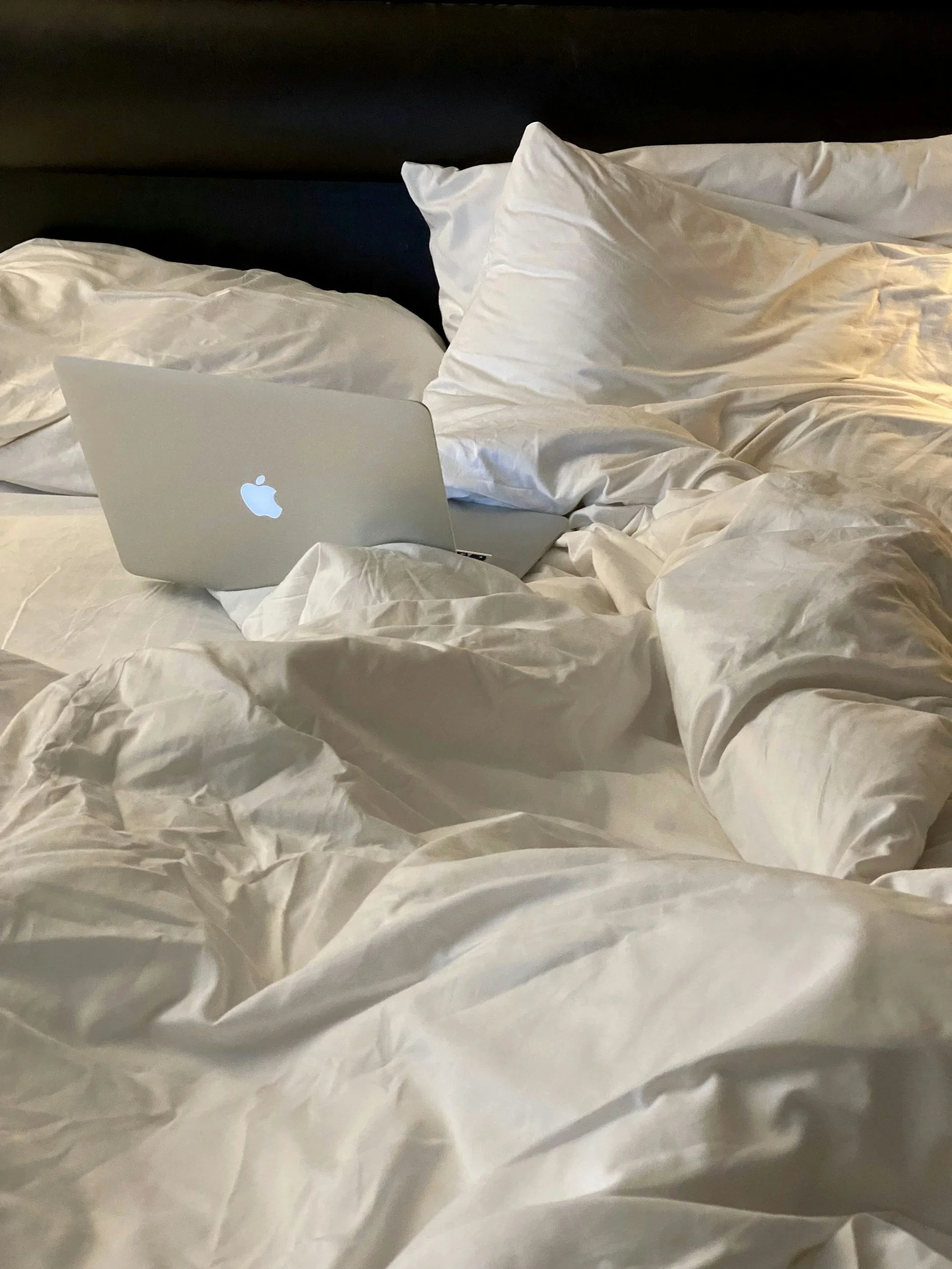 A laptop on a rumpled white bed with several white pillows and a dark headboard.