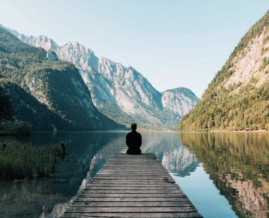 Person sitting on a wooden dock by a calm lake surrounded by mountains and forested hills.