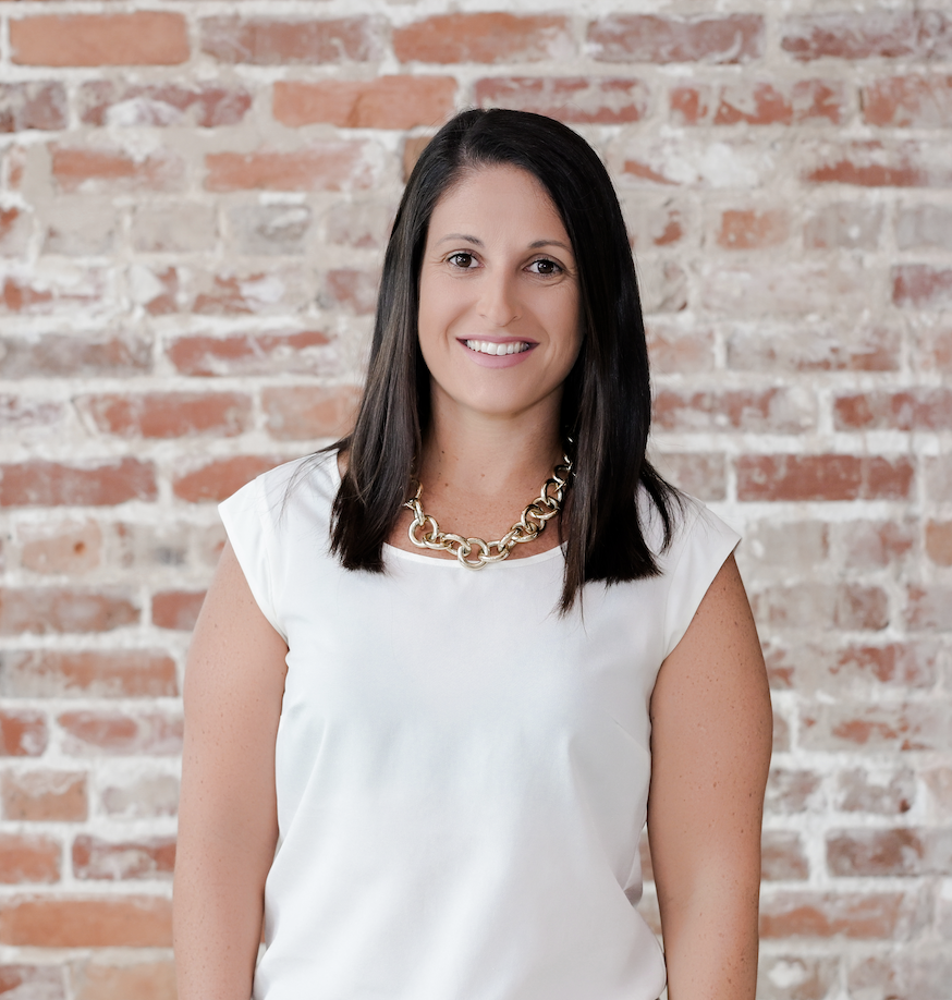 A woman with shoulder-length dark hair, wearing a white top and a chunky gold necklace, standing in front of a brick wall and smiling.