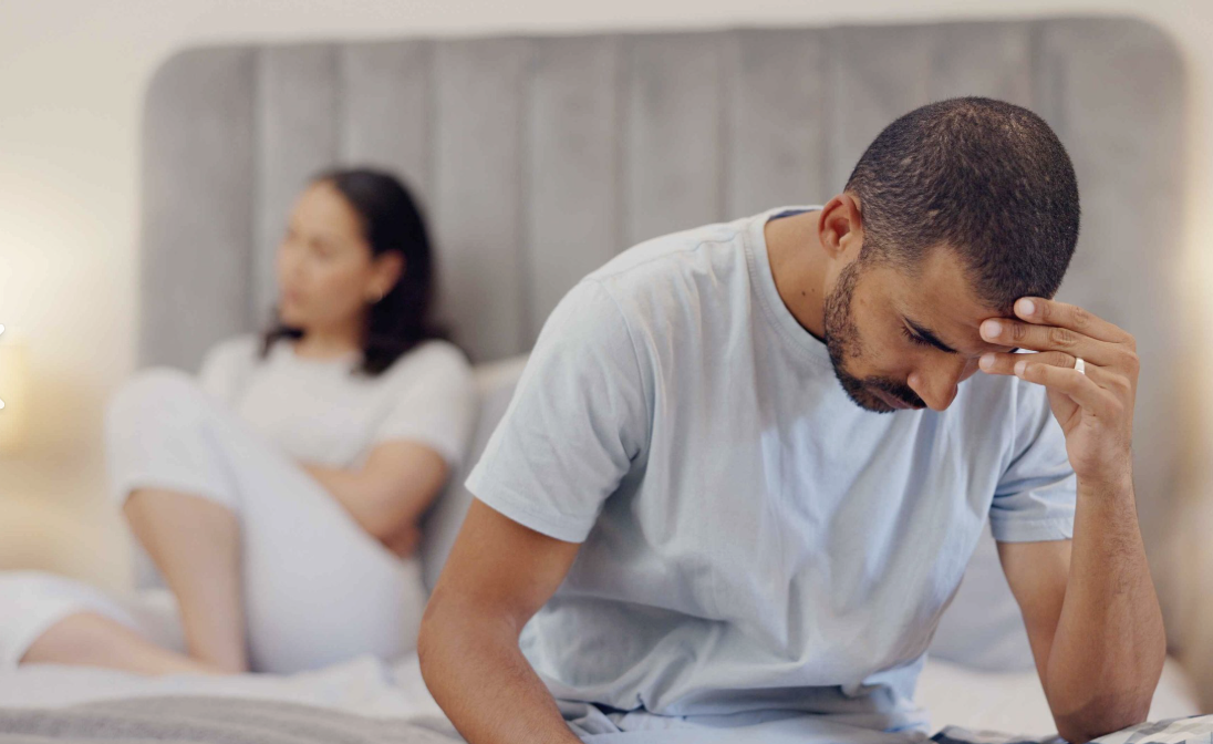 A distressed man sitting on a bed holding his forehead, with a woman sitting in the background.