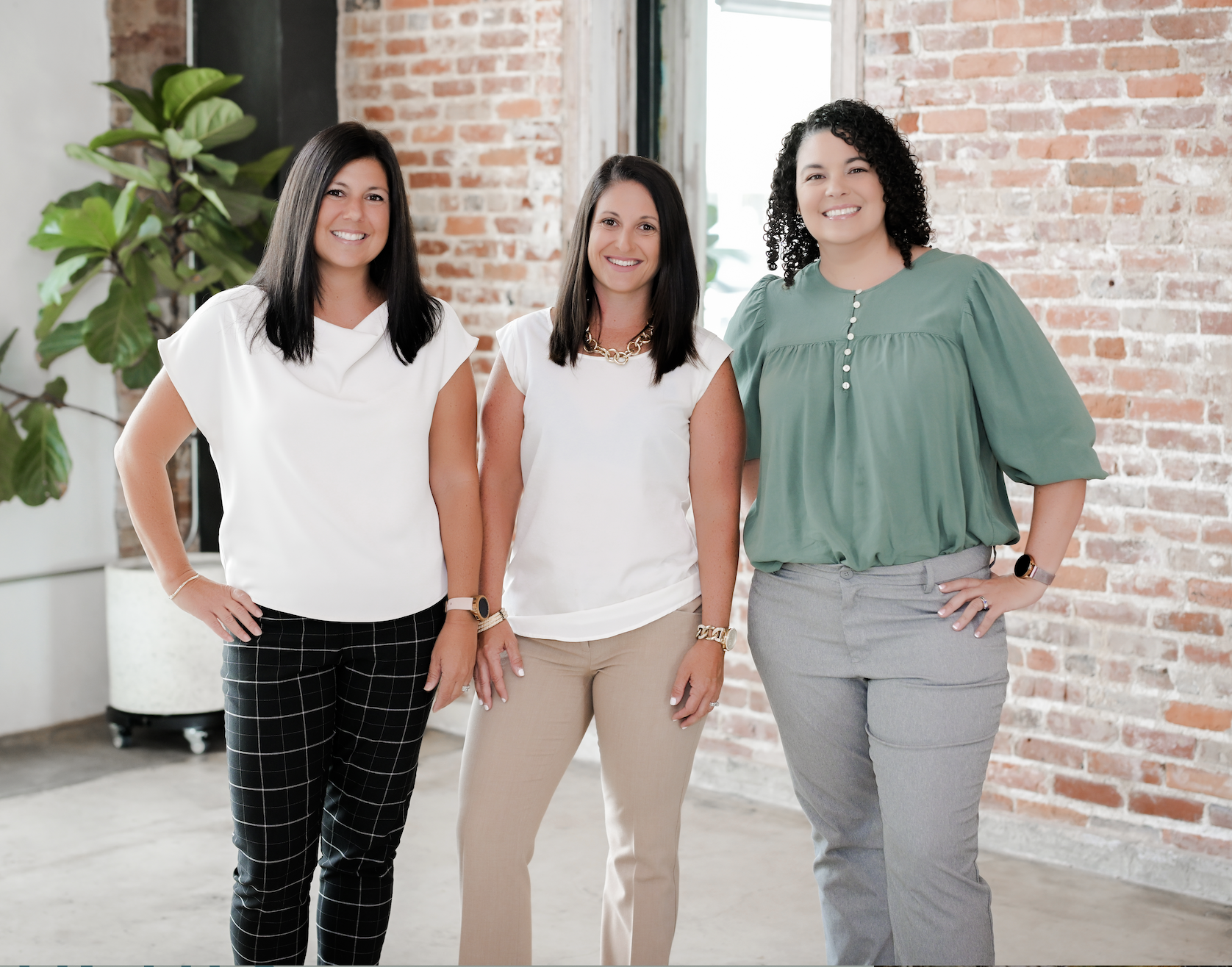 Three women standing indoors against a brick wall, smiling at the camera.