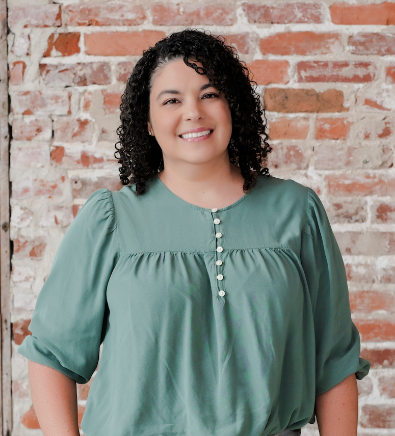 Smiling woman with curly black hair wearing a green blouse with pearl buttons, standing in front of a red brick wall.