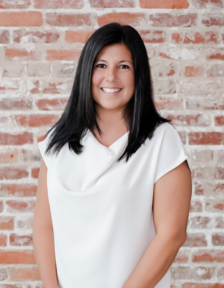 A woman with shoulder-length black hair, smiling, wearing a white top, standing in front of a brick wall.