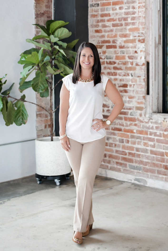 Woman in white sleeveless top and beige pants standing indoors near a large leafy plant and brick wall, smiling at the camera.