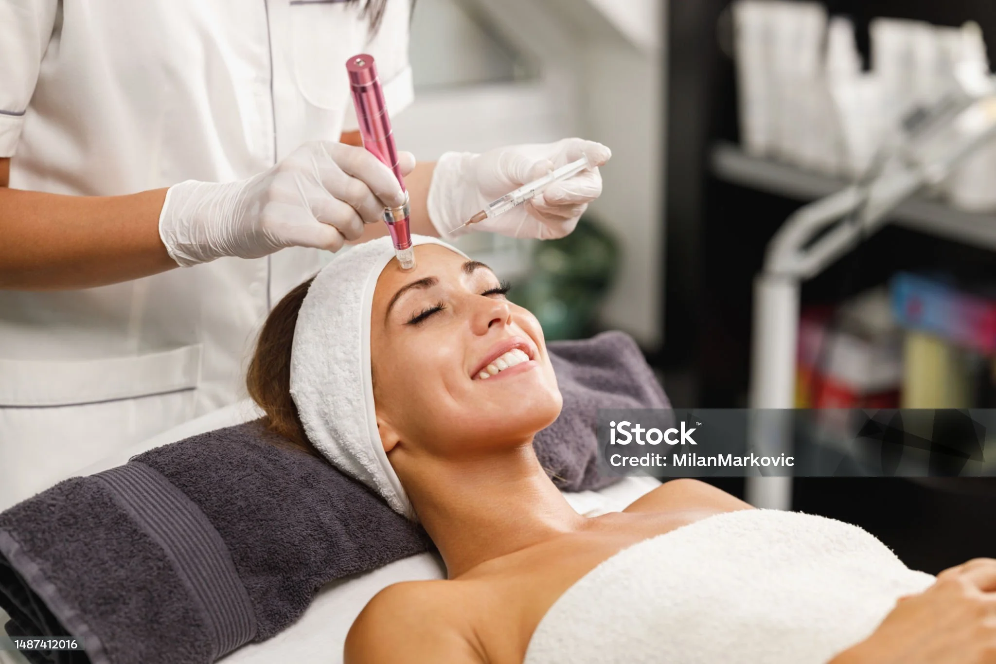 A woman receiving a facial treatment in a spa or beauty clinic, smiling with her eyes closed while a professional administers a cosmetic procedure.
