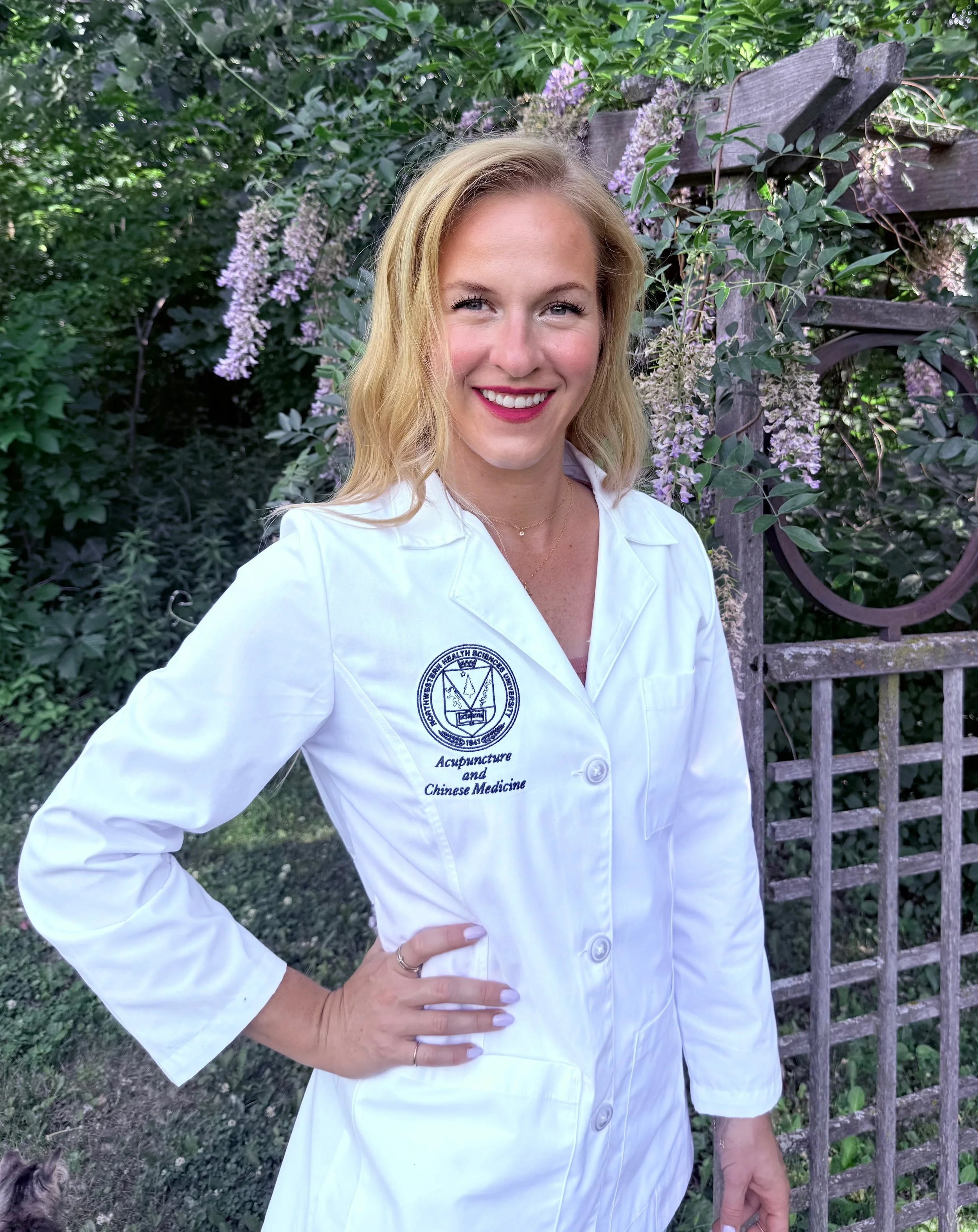 A woman with blonde hair wearing a white lab coat with embroidered text standing outdoors near greenery and purple flowers.