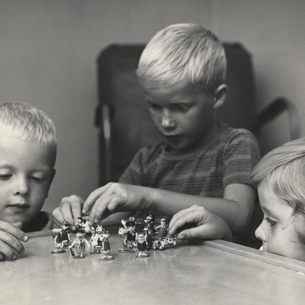 Black and white photo of Steve Pilcher playing with Hanna-Barbera “TV-Tinykins” by MARX toys, on a glass tabletop, flanked by his younger brother and sister.
