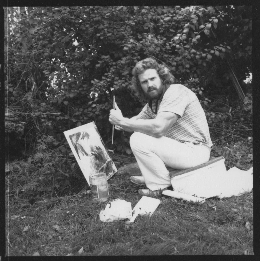 Black and white photo of Steve Pilcher painting outdoors holding a brush in front of an art panel in a forest setting.