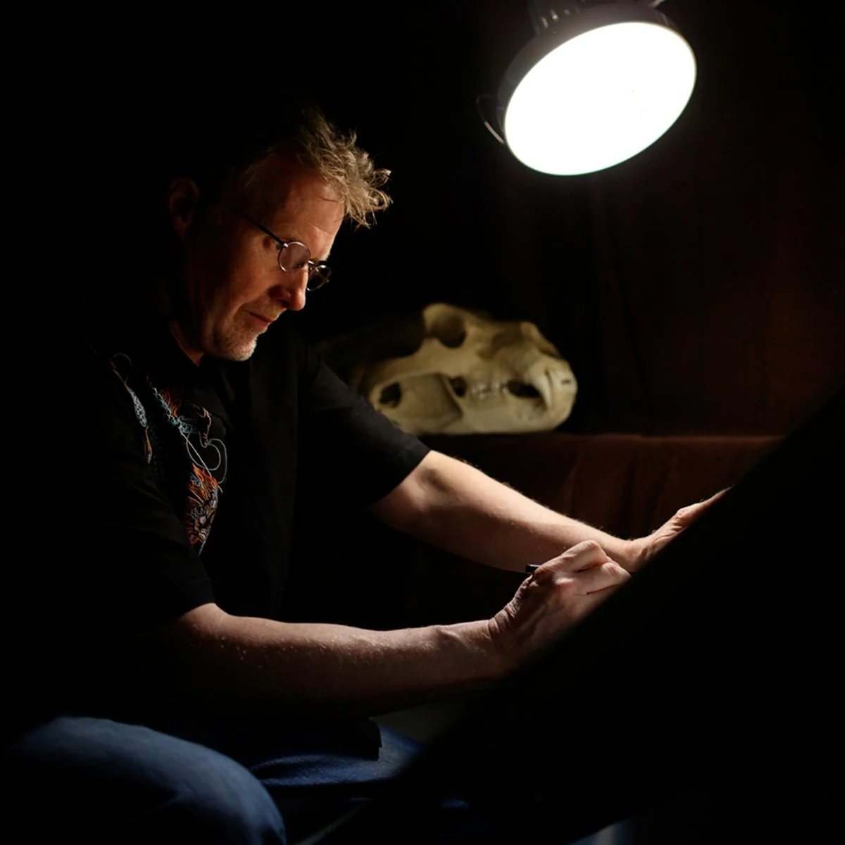 Photo of Steve Pilcher drawing at a desk lit by a lamp, with a bear skull in the background, photo by Deborah Coleman.