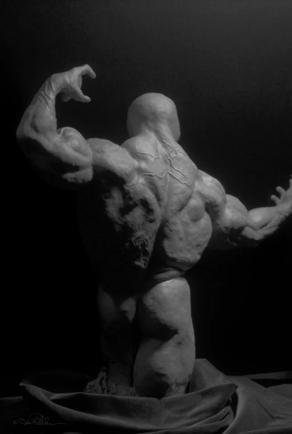Sculpture in grey clay of muscular body builder from the back with one arm extended and the other arm flexed up high.