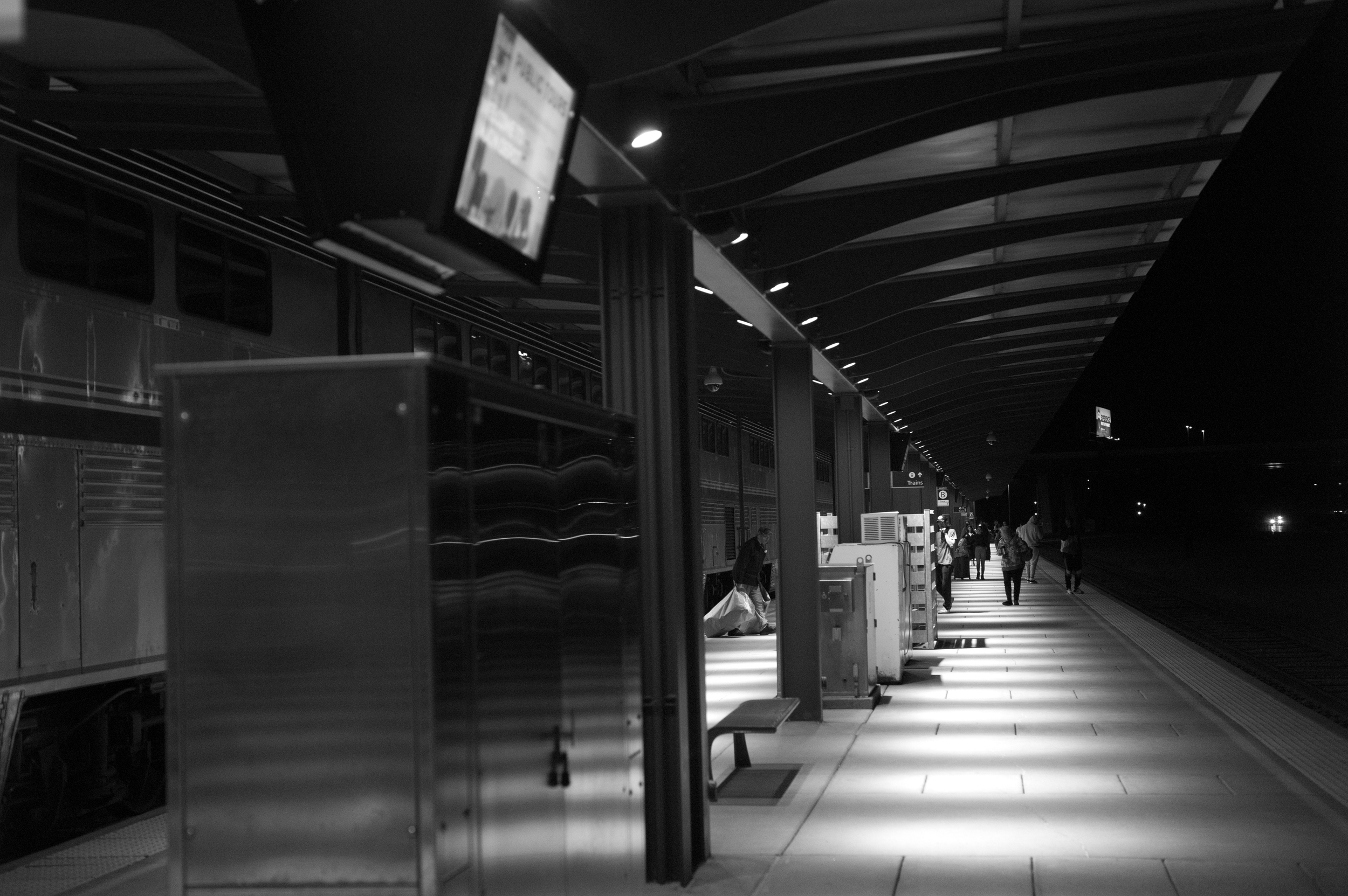 Nighttime scene at a train station platform with a train on the left and a few passengers walking or waiting.