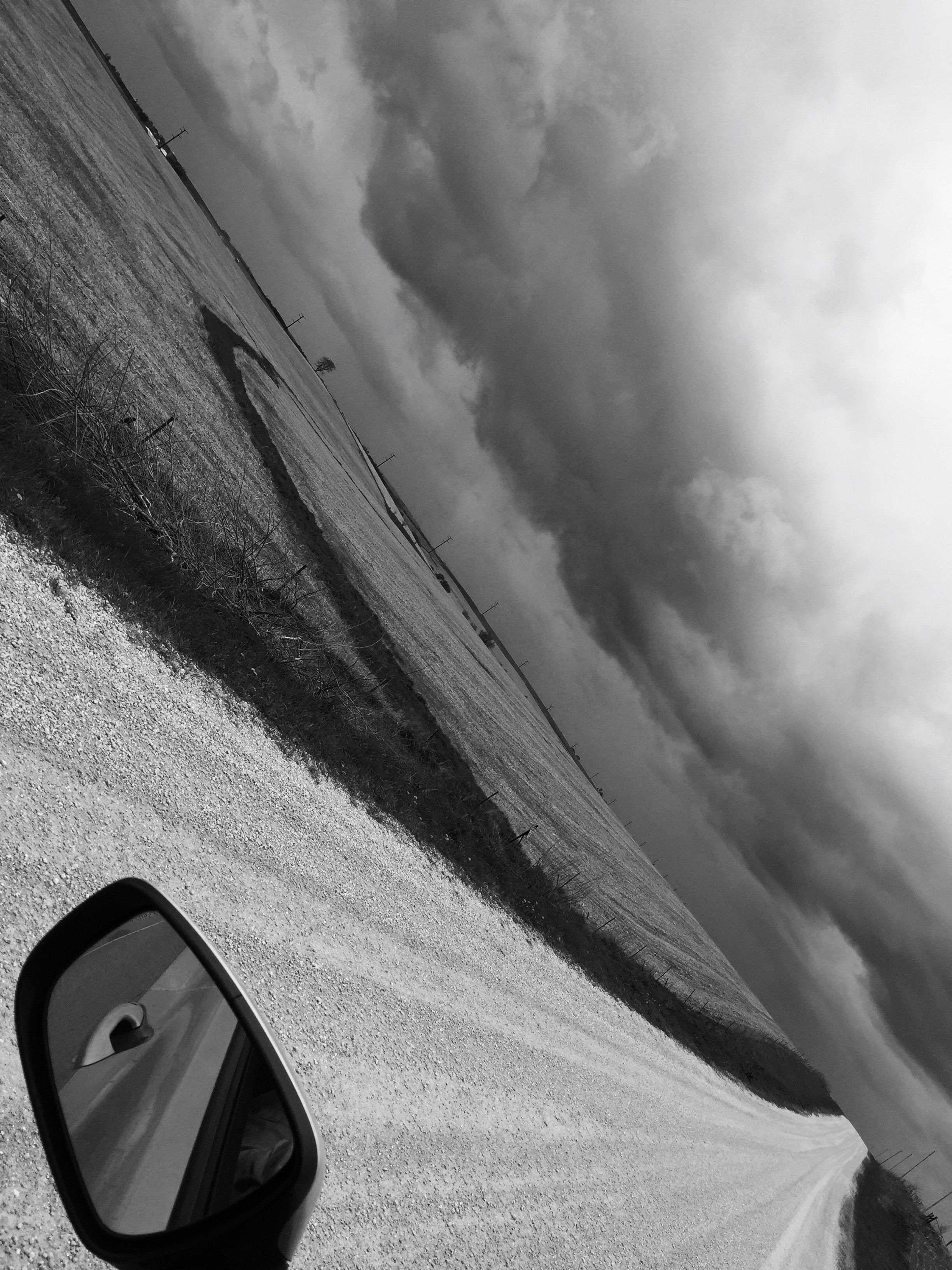 Black and white photo of a rural gravel road stretching into the distance with storm clouds overhead. A car's side mirror is visible in the lower left corner.