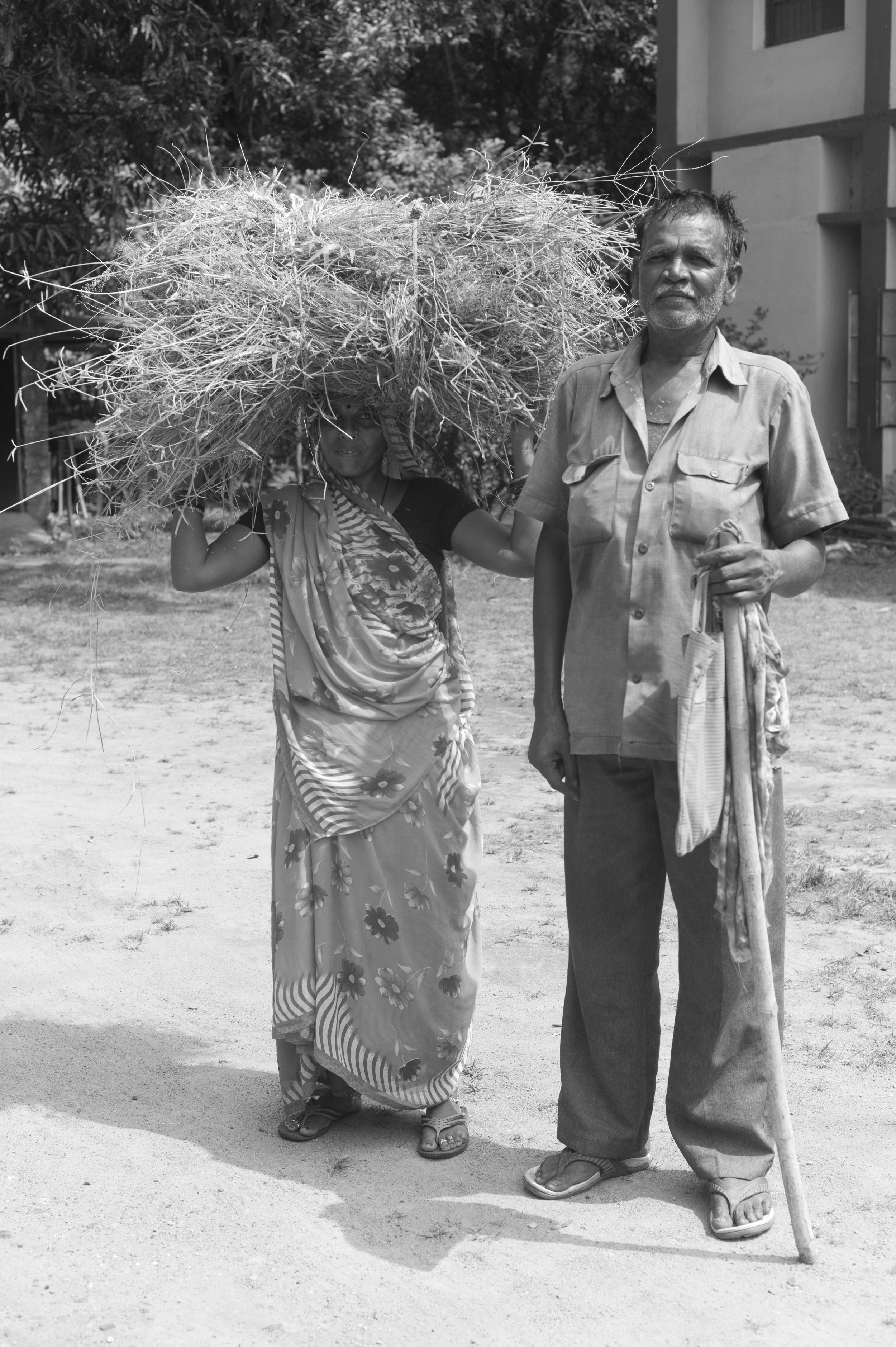 A black and white photo of an elderly man standing next to a woman with a large bundle of dry straw on her head, outdoors on a dirt ground with trees and a building in the background.