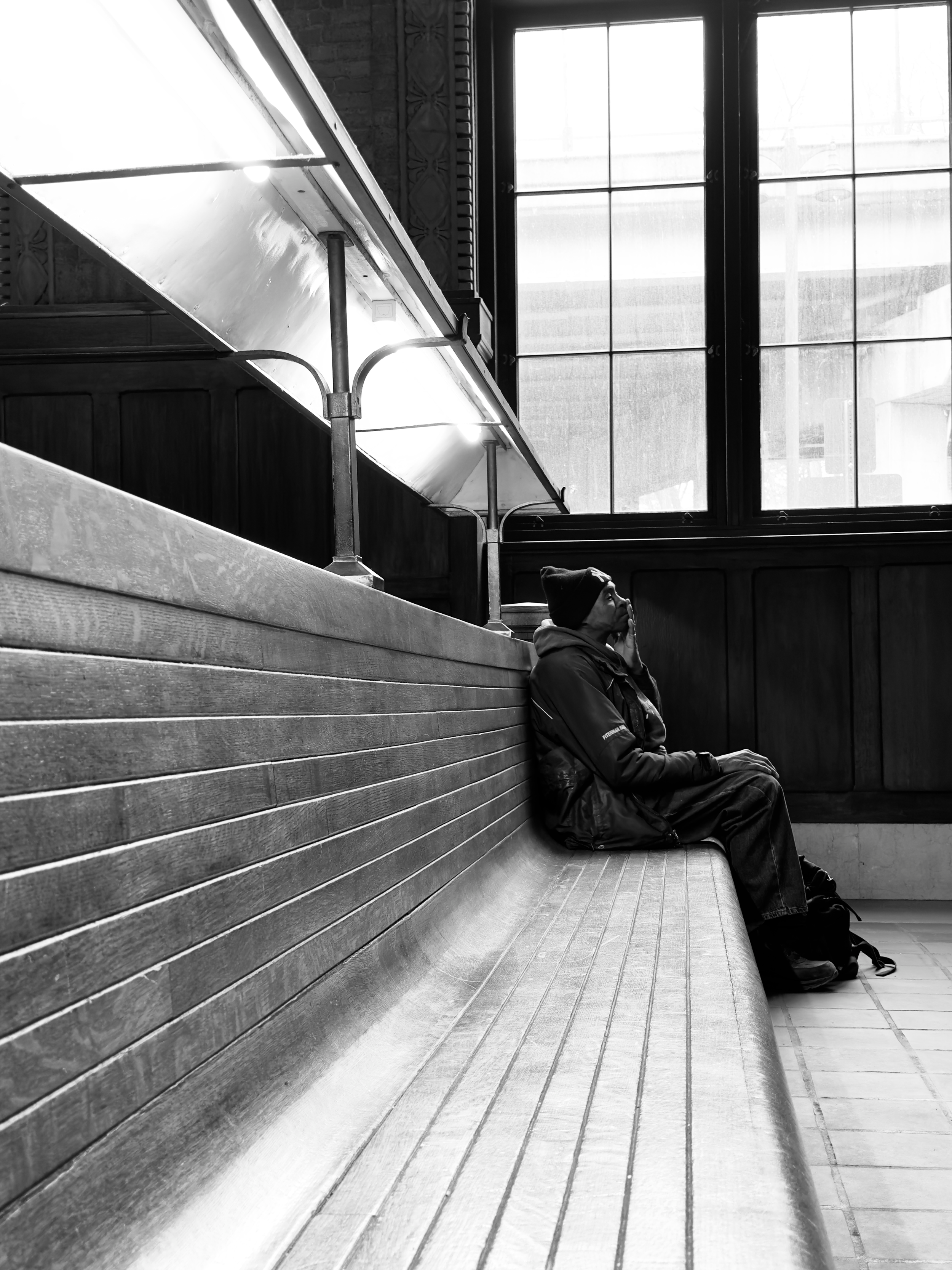 A man sitting alone on a wooden bench inside a building with large windows, wearing a hat and a jacket, possibly waiting or in thought, in black and white.